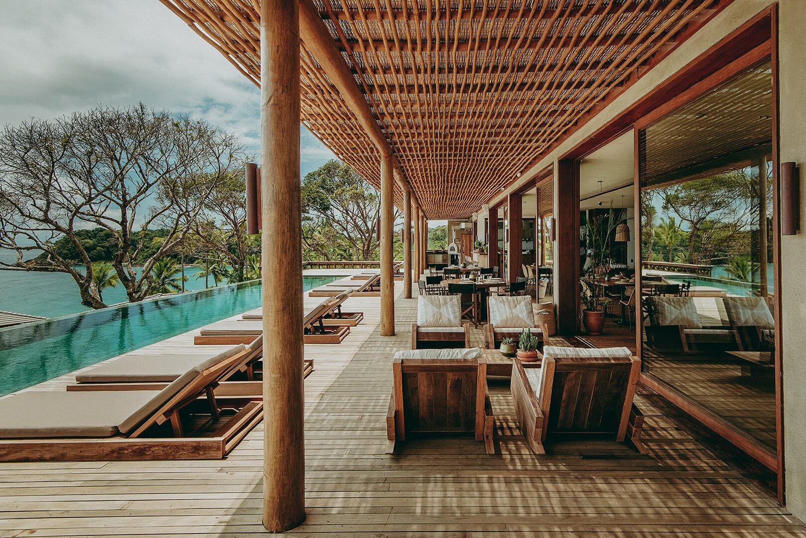Barracuda Hotel & Villas, South America — Waterfront terrace with teak lounge chairs, infinity pool, wooden lattice pergola, and floor-to-ceiling glass doors overlooking tropical landscape.