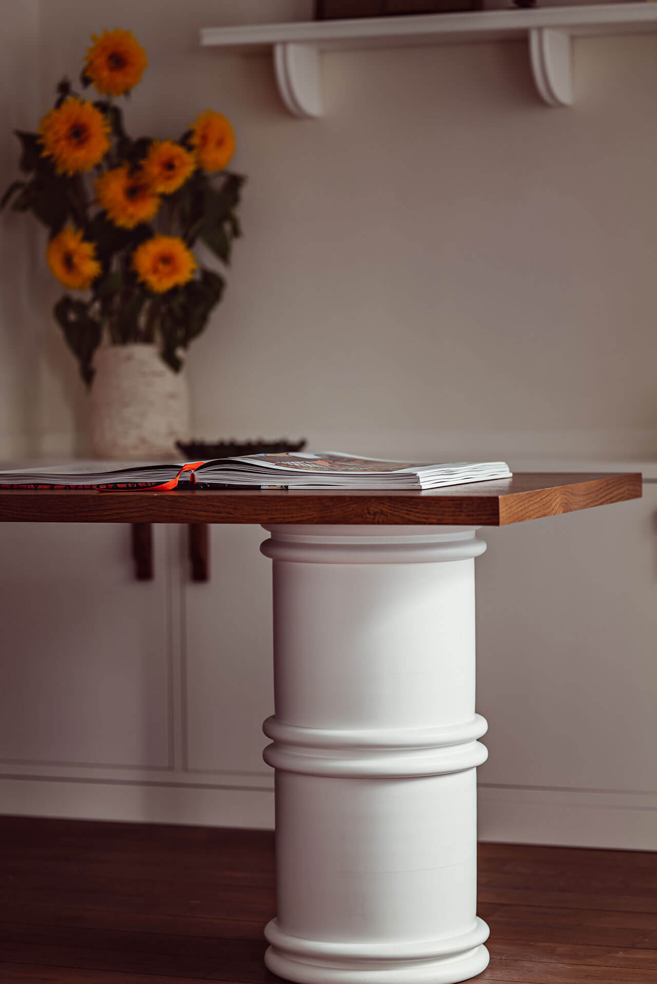 Académies et des Arts, Europe — Minimalist pedestal table with wood top, open magazine, sunflowers in white vase, neutral interior architecture.