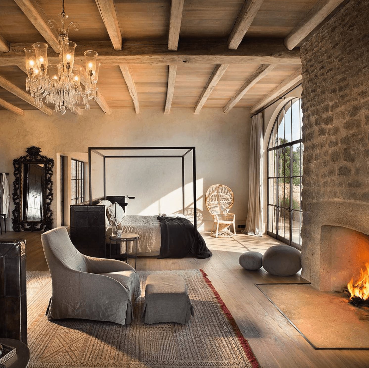 Arijiju Retreat, Africa — Sunlit bedroom with exposed wooden beams, stone walls, fireplace, canopy bed, wicker chair, arched glass doors opening to landscape.