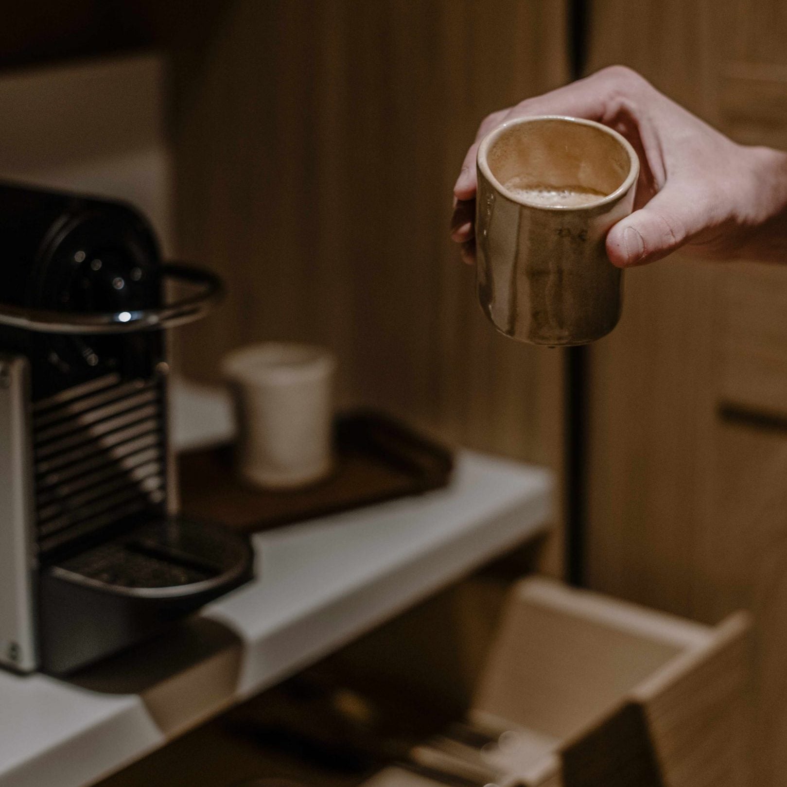 Casa di Langa, Europe — Hand holding ceramic cup of espresso beside black coffee machine, warm interior lighting, minimalist hospitality design.