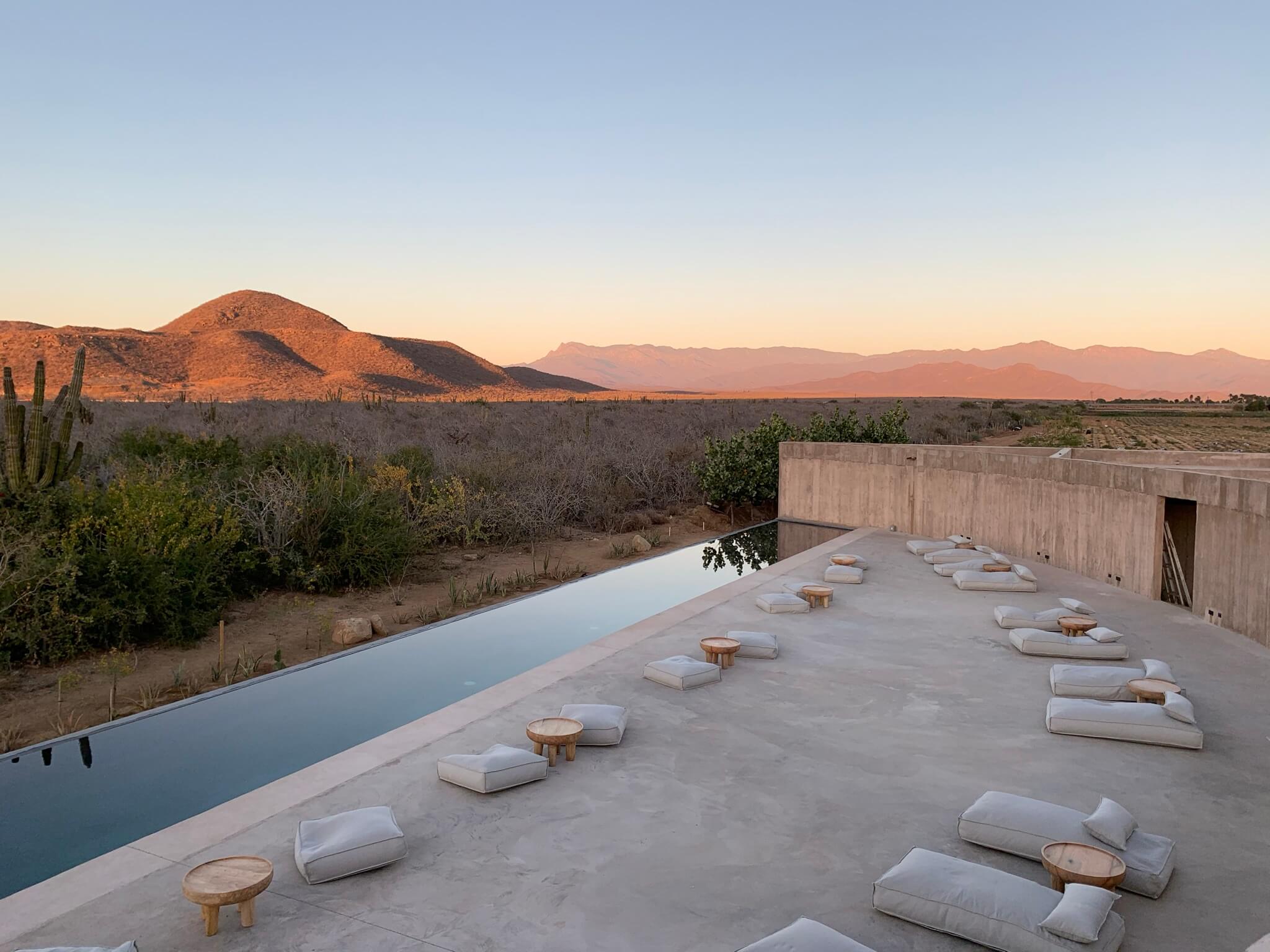 Paradero Todos Santos, Mexico — Infinity pool and minimalist loungers overlooking desert mountains at sunset, concrete terrace with wooden accents and saguaro cactus.