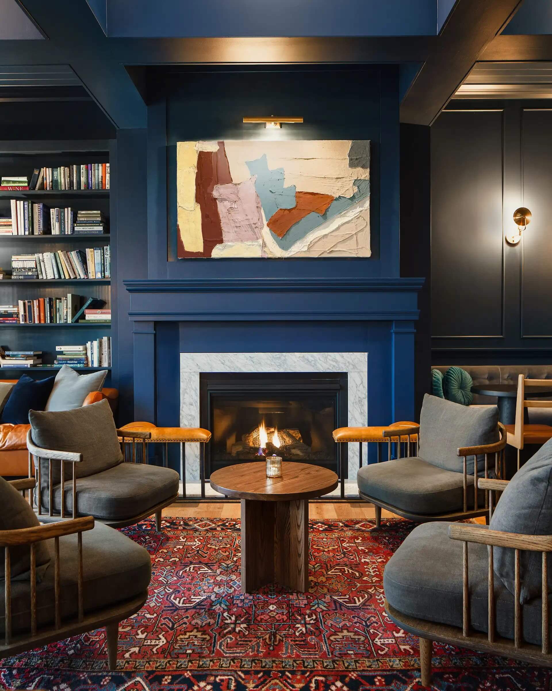 The Lake House, North America — Contemporary living room with deep blue walls, marble fireplace, mid-century wooden chairs, abstract artwork, built-in bookshelves, and glowing brass sconces.