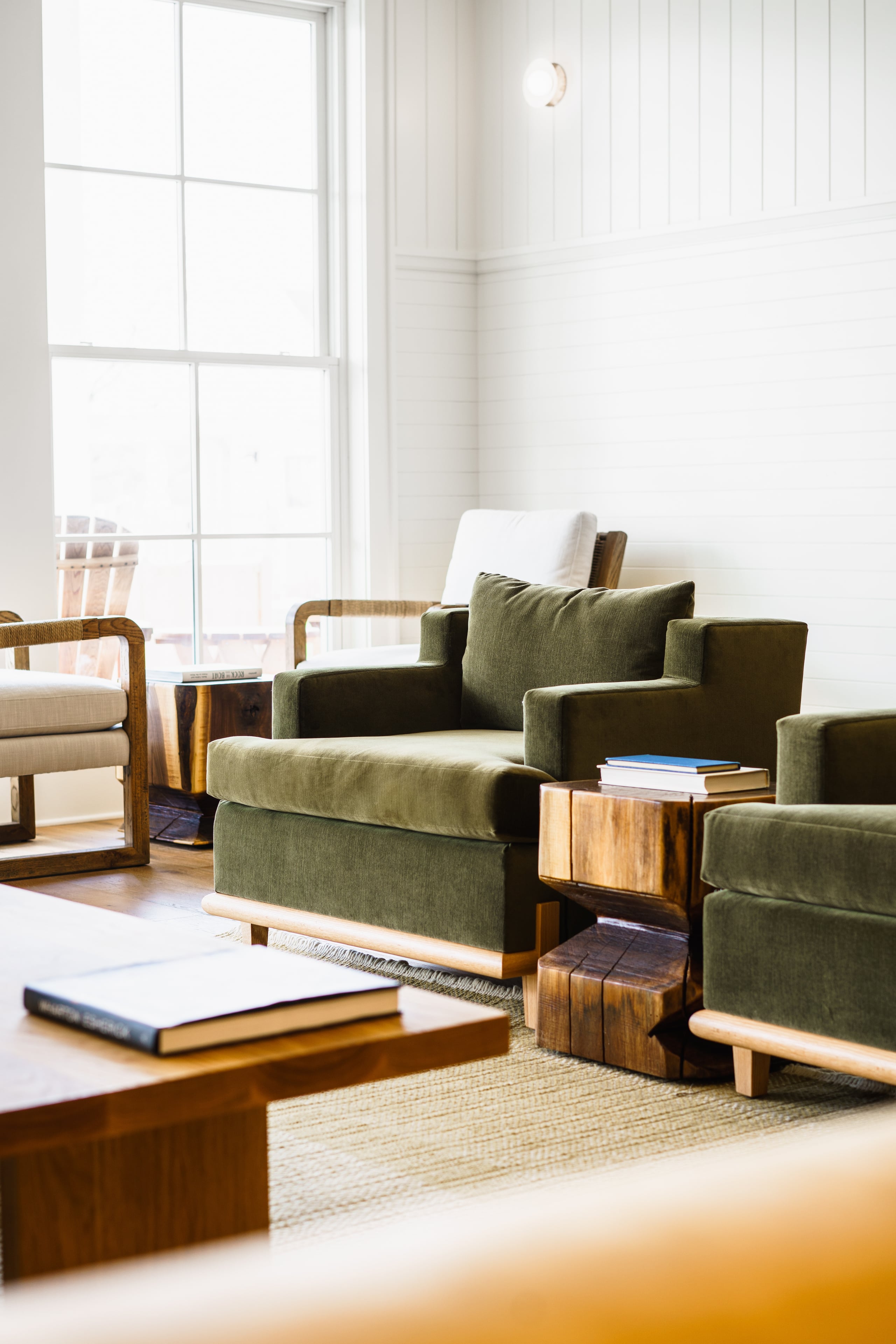 The Lake House, North America — Sunlit living room with sage green upholstered armchairs, walnut side tables, natural fiber rug, and floor-to-ceiling windows overlooking water.