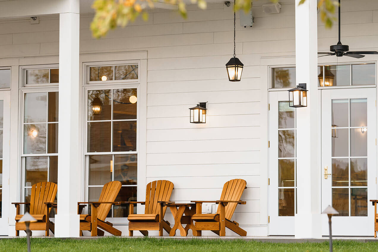 The Lake House, North America — Modern farmhouse porch with white shiplap, golden Adirondack chairs, pendant lanterns, and floor-to-ceiling windows at dusk.