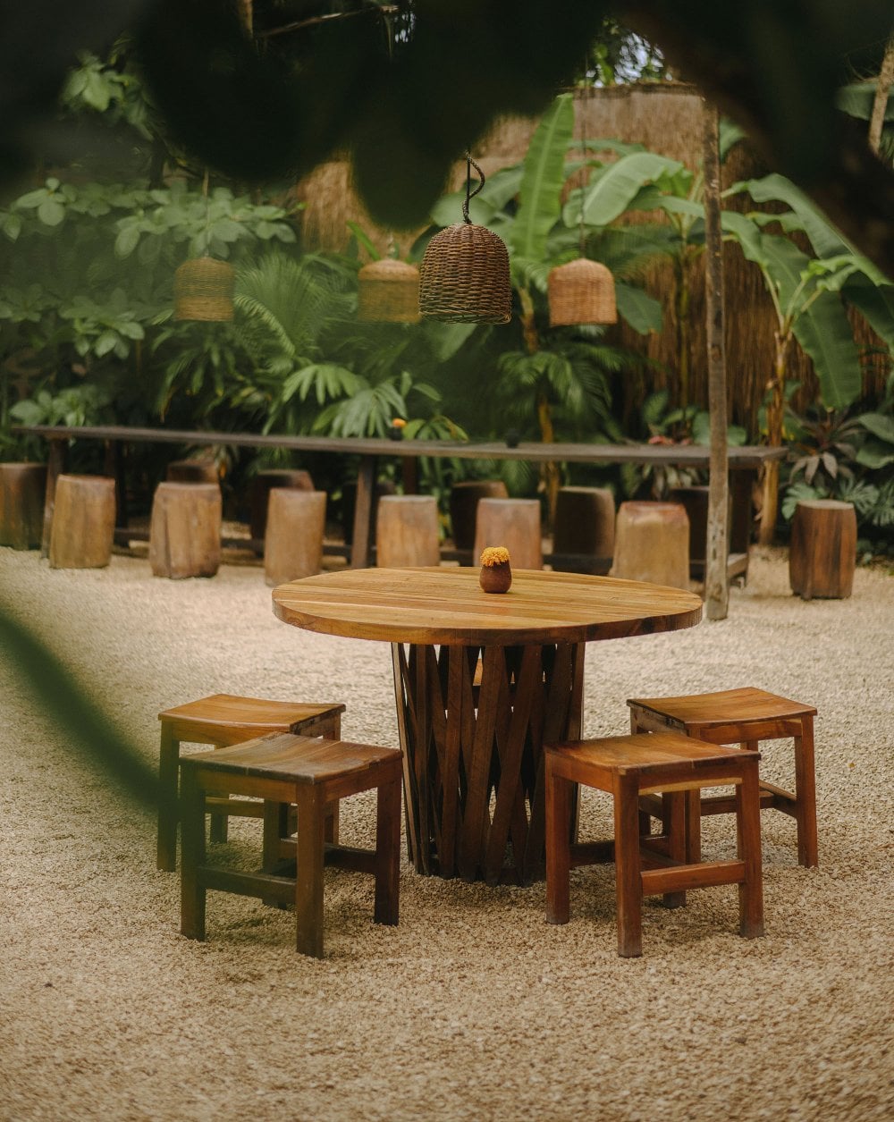Tulum Treehouse, Mexico — Round wooden table with woven pendant lamp and log stools on sand, surrounded by lush tropical foliage and wooden pavilion.