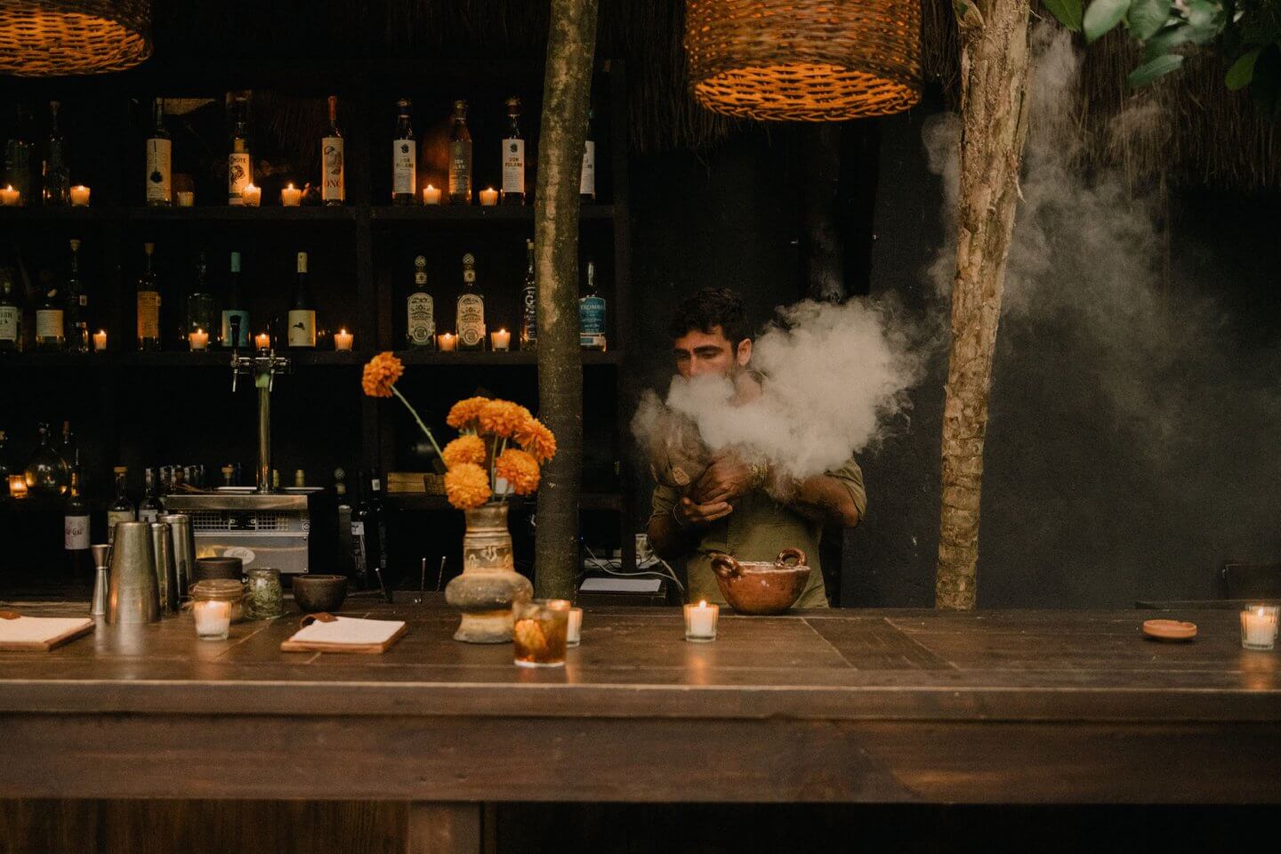 Tulum Treehouse, Mexico — Candlelit bar interior with dark wood counter, amber flowers, bottle-lined shelves, and bartender creating dramatic dry ice cocktail smoke.