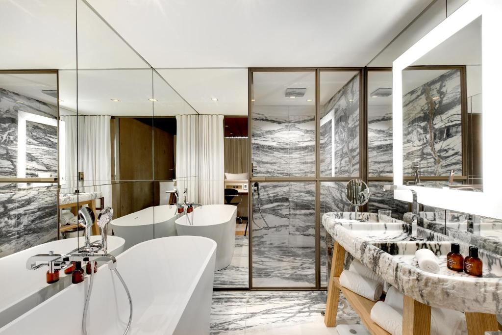 Lily of the Valley, Europe — Luxe bathroom with marble-veined walls, freestanding soaking tub, dual vanities, frameless glass shower enclosure, warm wood accents, and integrated lighting.