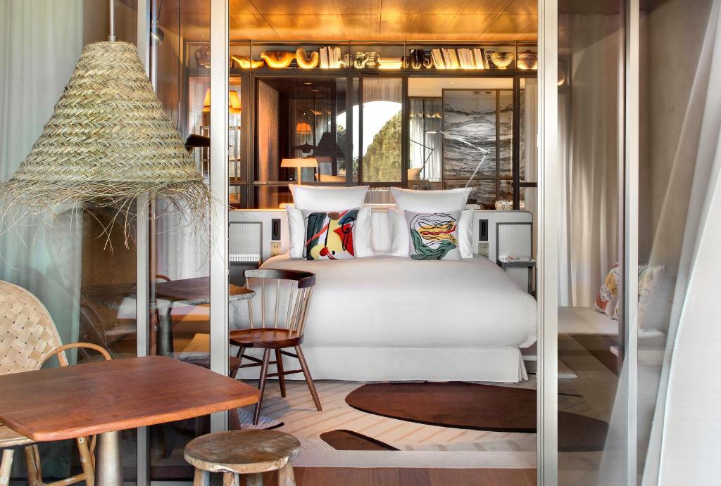 Lily of the Valley, Europe — Modern hotel bedroom with woven pendant lamp, colorful abstract pillows, wooden furniture, and glass partition opening to lounge area.