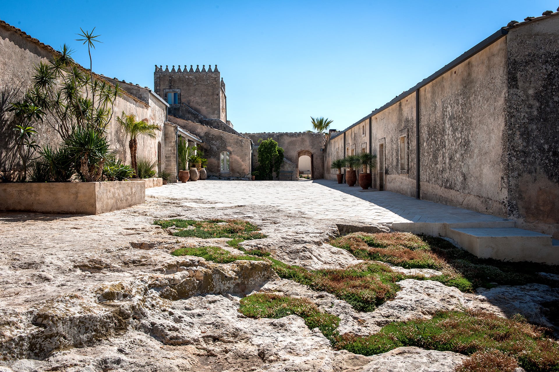 Dimora delle Balze, Europe — Sunlit courtyard with weathered stone walls, crenellated tower, terracotta planters, and Mediterranean architectural details.