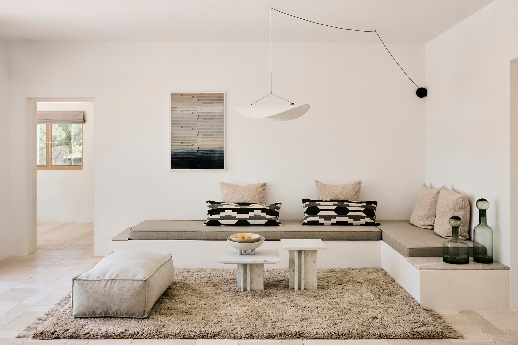 Villa Sessana, Italy — Minimalist living room with built-in seating, geometric patterned pillows, travertine tables, neutral textiles, and sculptural pendant light suspended above.