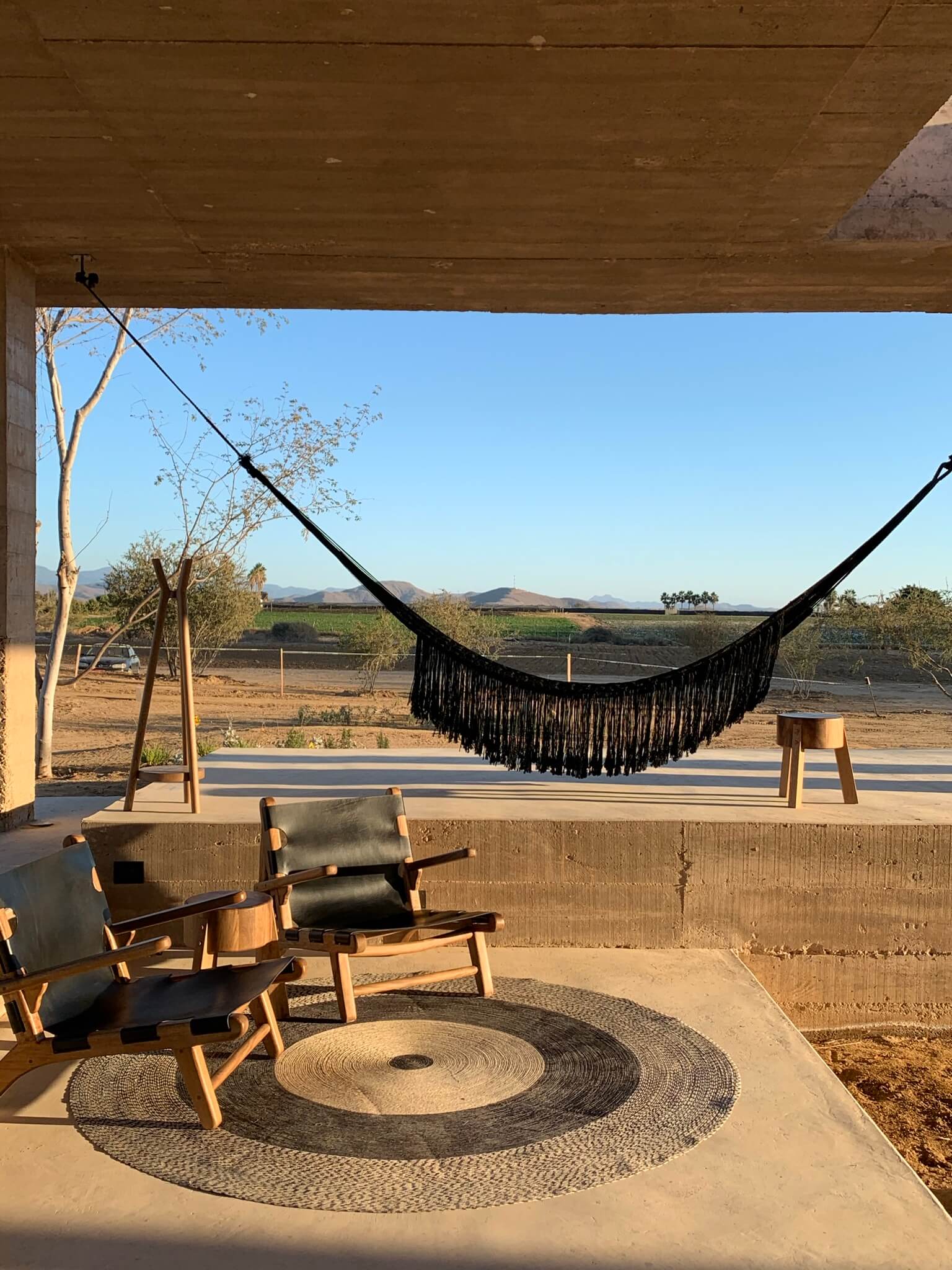 Paradero Todos Santos, Mexico — Covered pavilion with black fringed hammock, wooden lounge chairs, spiral rope rug, and agricultural valley views at sunset.
