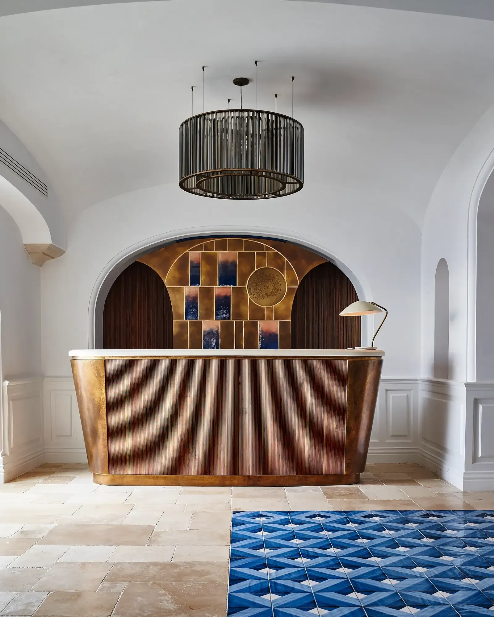 Borgo Santandrea, Europe — Bronze reception desk with ribbed detailing beneath arched niche featuring golden shelving and blue patterned floor tiles in minimalist white corridor.