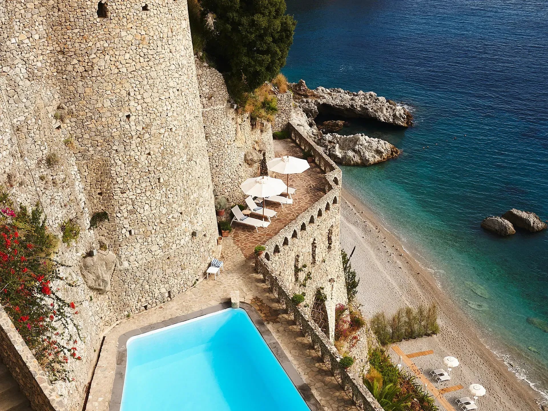 Borgo Santandrea, Europe — Clifftop pool and terraced beach with stone fortifications, azure Mediterranean waters, white umbrellas, and blooming bougainvillea.