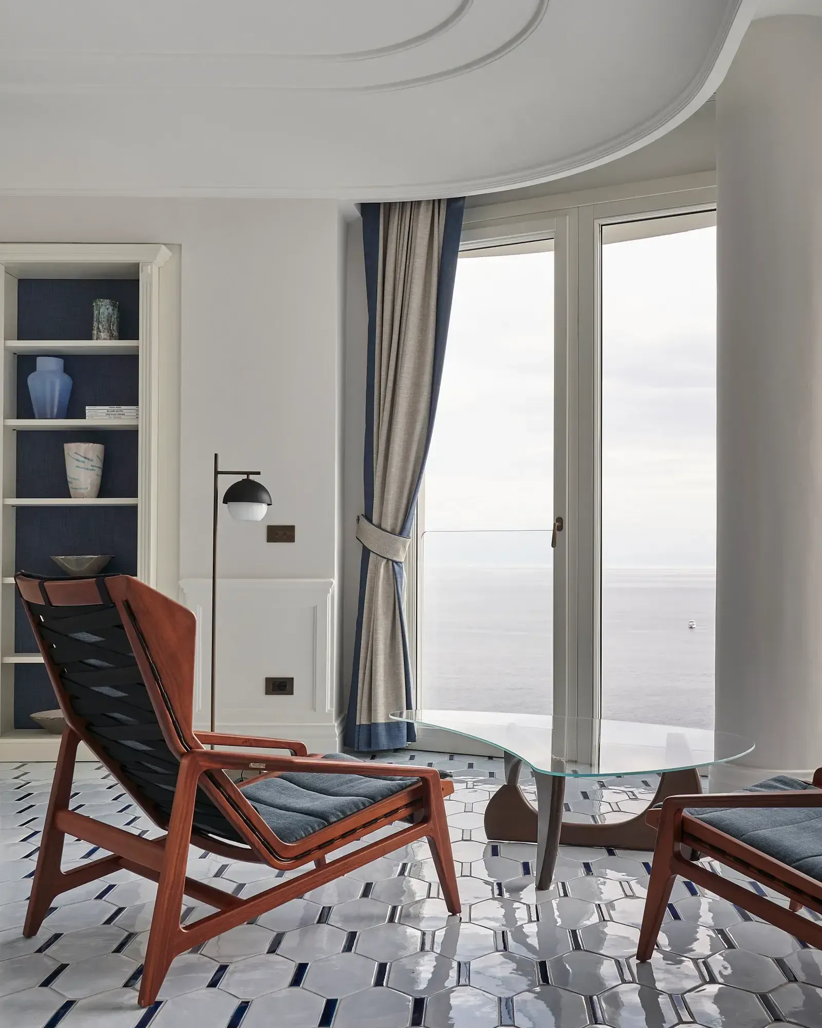 Borgo Santandrea, Europe — Curved ceiling detail with mid-century lounge chairs, glass table, floor-to-ceiling windows overlooking coastline, navy built-in shelving, geometric tile flooring.