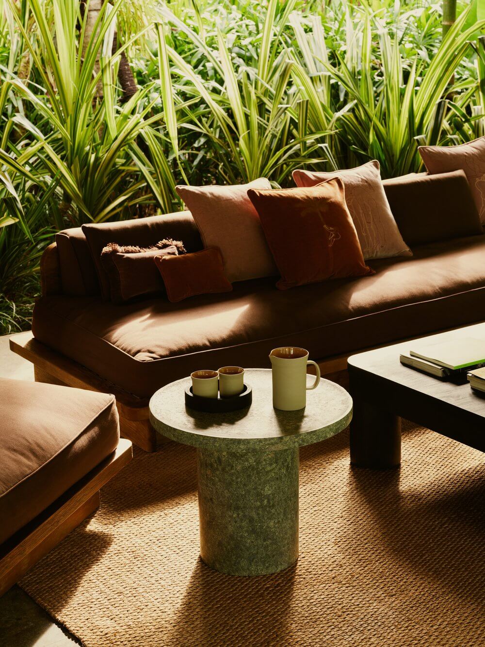 Lost Lindenberg, Asia — Brown linen sofa with rust pillows beside striped green plants, green stone side table with ceramic vessels, natural light casting shadows on jute rug.