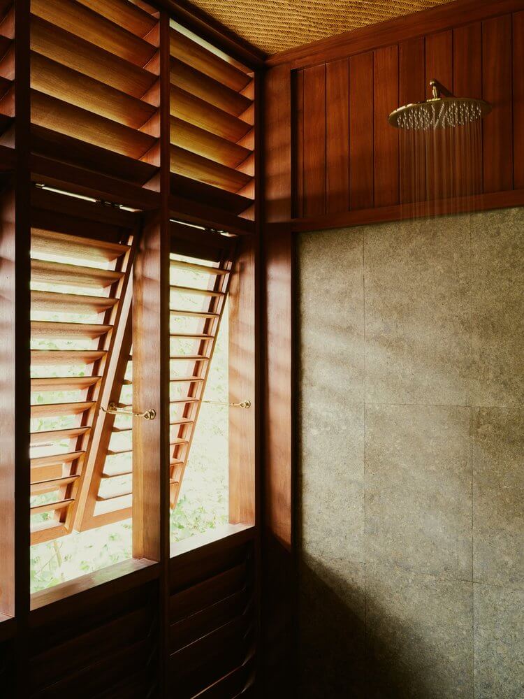 Lost Lindenberg, Asia — Wooden louvered shutters frame natural light in minimalist shower with stone tile walls and rainfall showerhead.