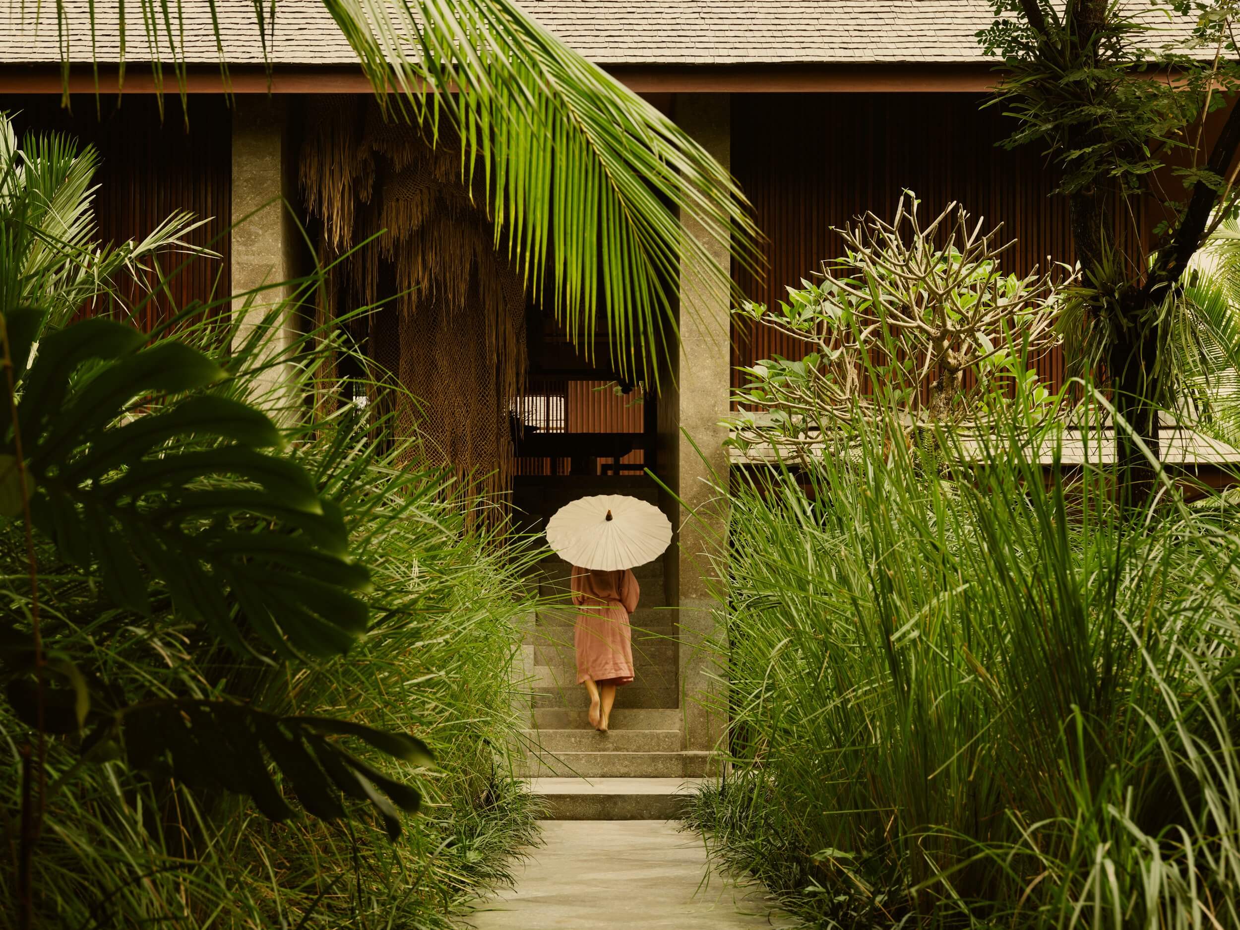 Lost Lindenberg, Asia — Woman with white parasol walking through lush tropical garden corridor toward modernist wooden villa with stone courtyard.