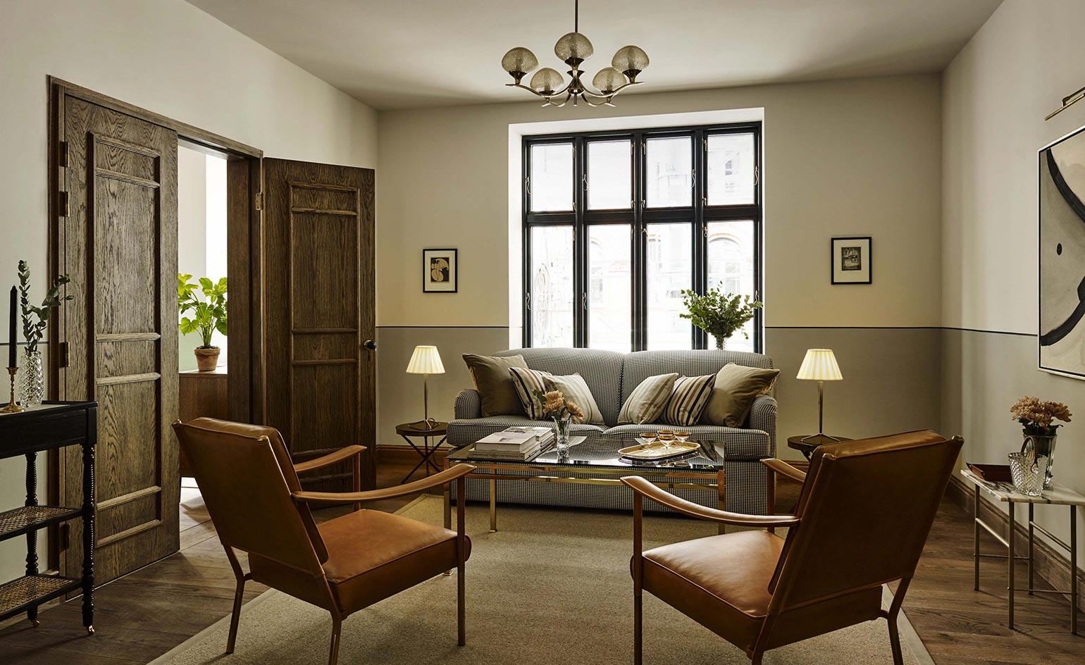 Hotel Sanders, Europe — Mid-century modern living room with cognac leather chairs, gray upholstered sofa, brass coffee table, dark wood paneling, and natural light from gridded windows.