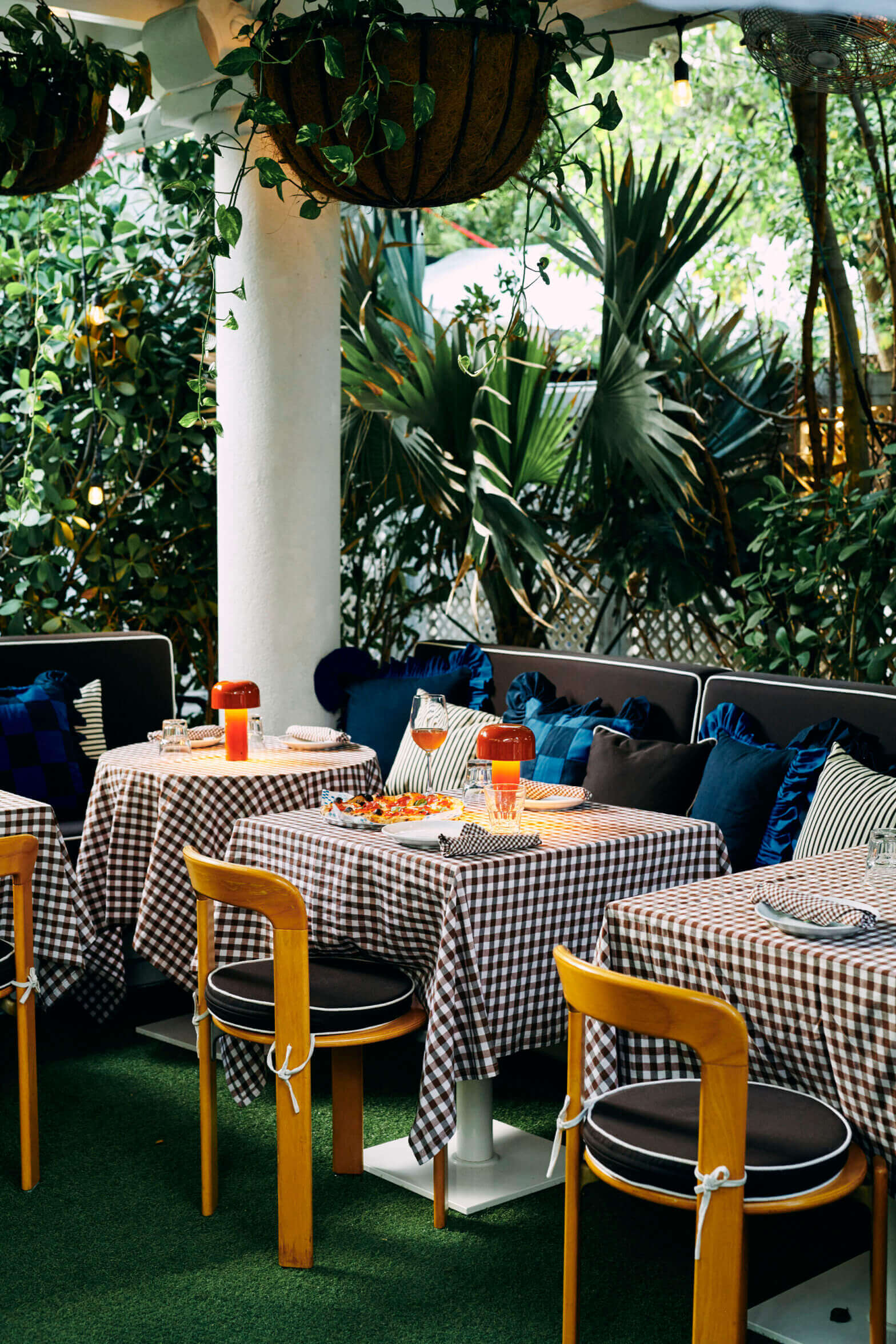 Paradise Pizza, North America — Lush tropical patio with gingham tablecloths, blue velvet seating, wooden chairs, hanging plants, and natural daylight filtering through verdant foliage.