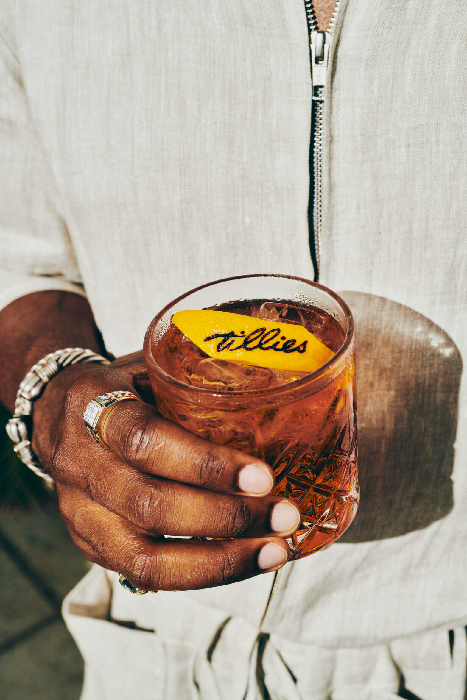 Palm Heights, North America — Hand holding Willies cocktail with yellow citrus peel garnish, cream linen jacket, rings, warm amber lighting.