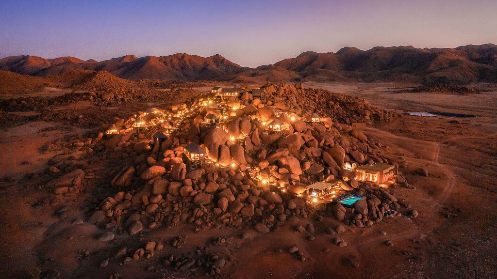 Zannier Sonop, Africa — Illuminated boulder-built luxury lodge nestled in desert landscape at dusk, warm ambient lighting, minimalist architecture integrated into natural rock formations.