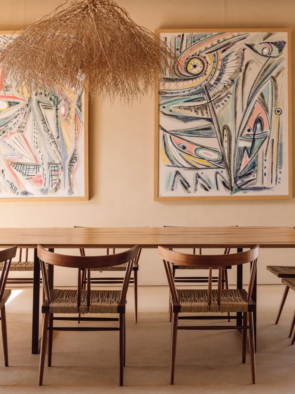 Melides Art, Portugal — Minimalist dining space with woven chairs, wooden table, abstract ink drawings on pale walls, dried grass pendant light fixture.