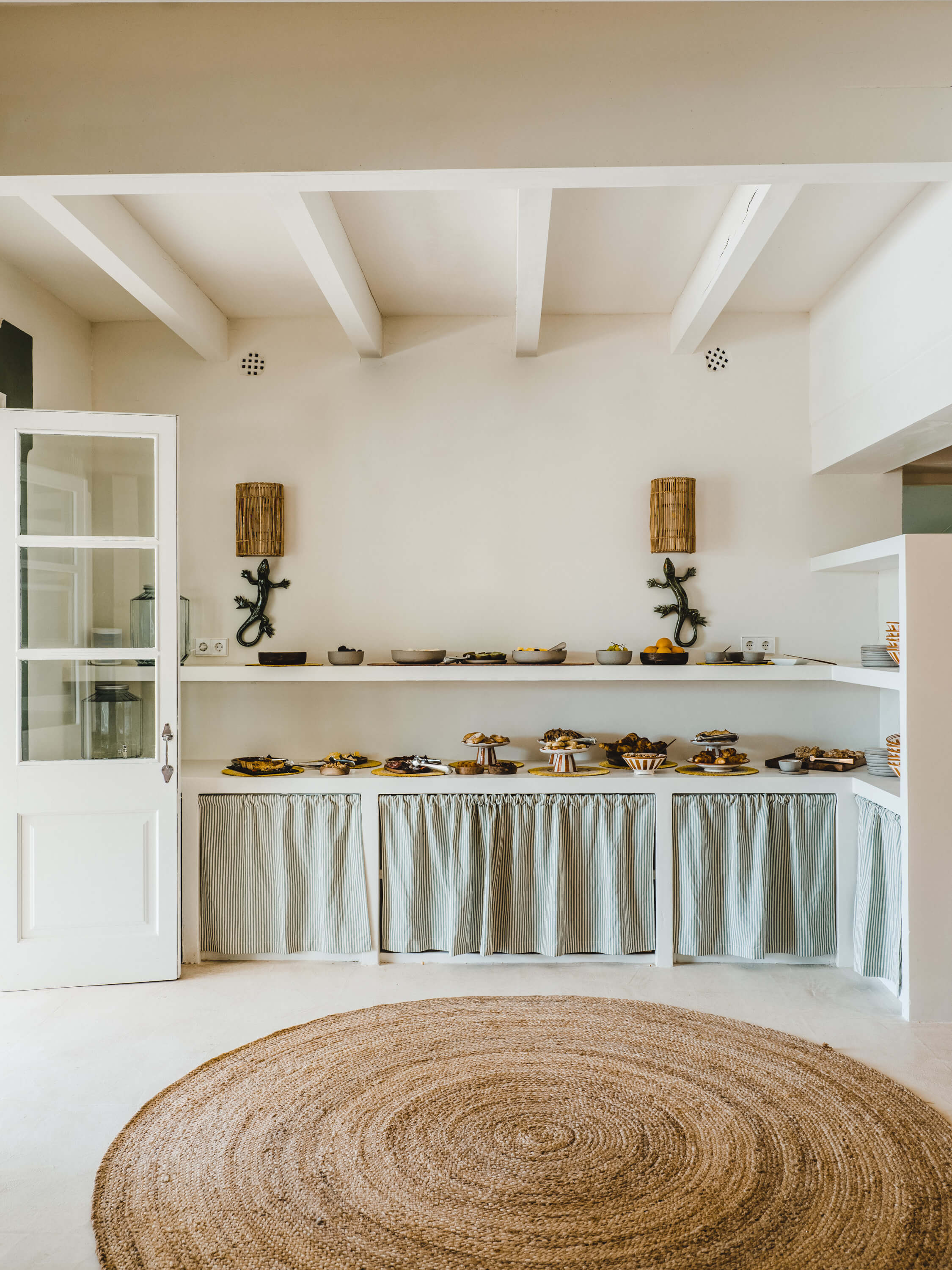 Morvedra Nou, Balearic Islands, Spain — Minimalist kitchen pantry with open shelving, striped linen curtains, exposed beam ceiling, and woven jute rug.