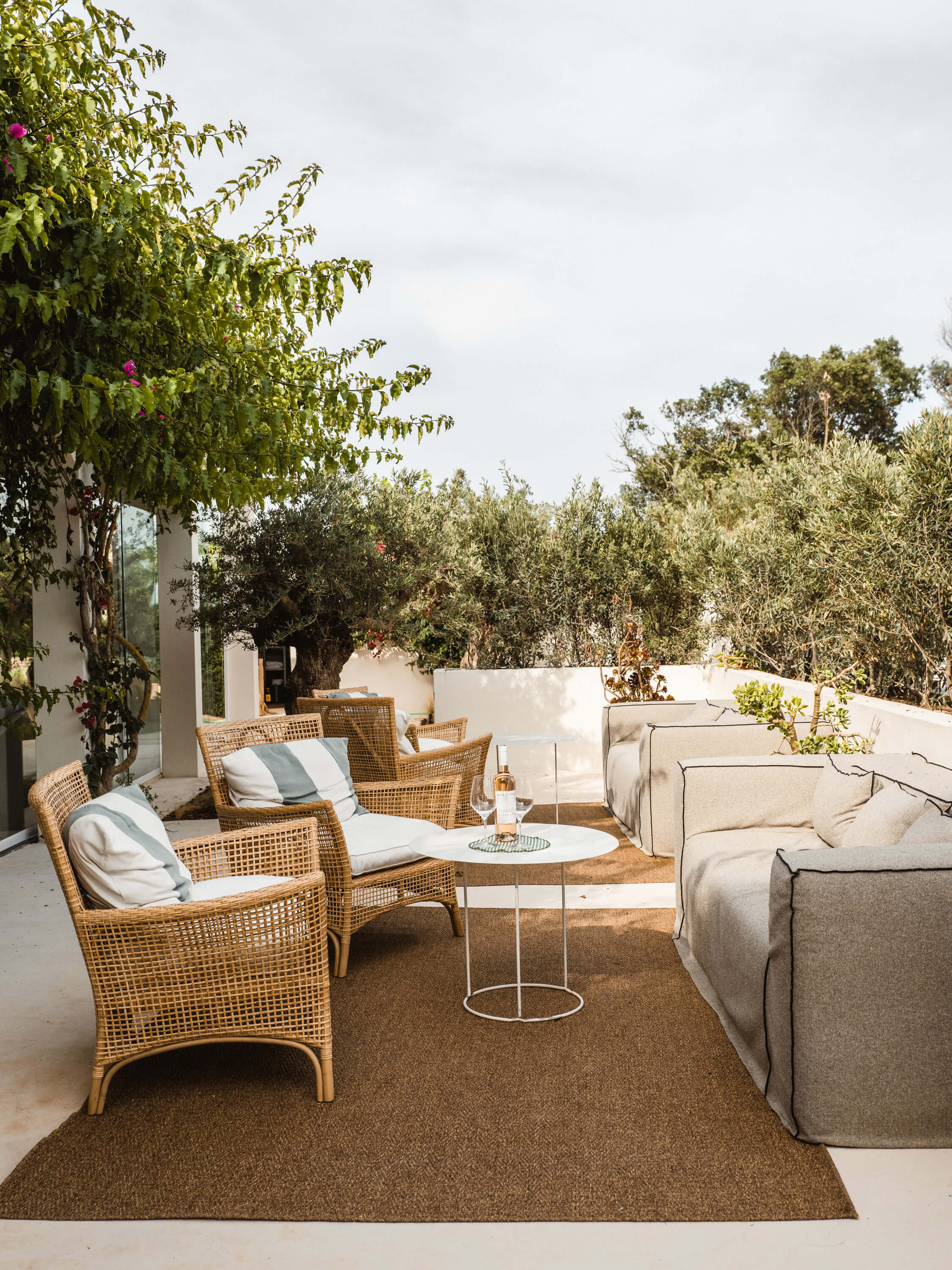 Morvedra Nou, Balearic Islands, Spain — Terrace seating area with woven rattan chairs, linen sofa, and olive trees under Mediterranean light.