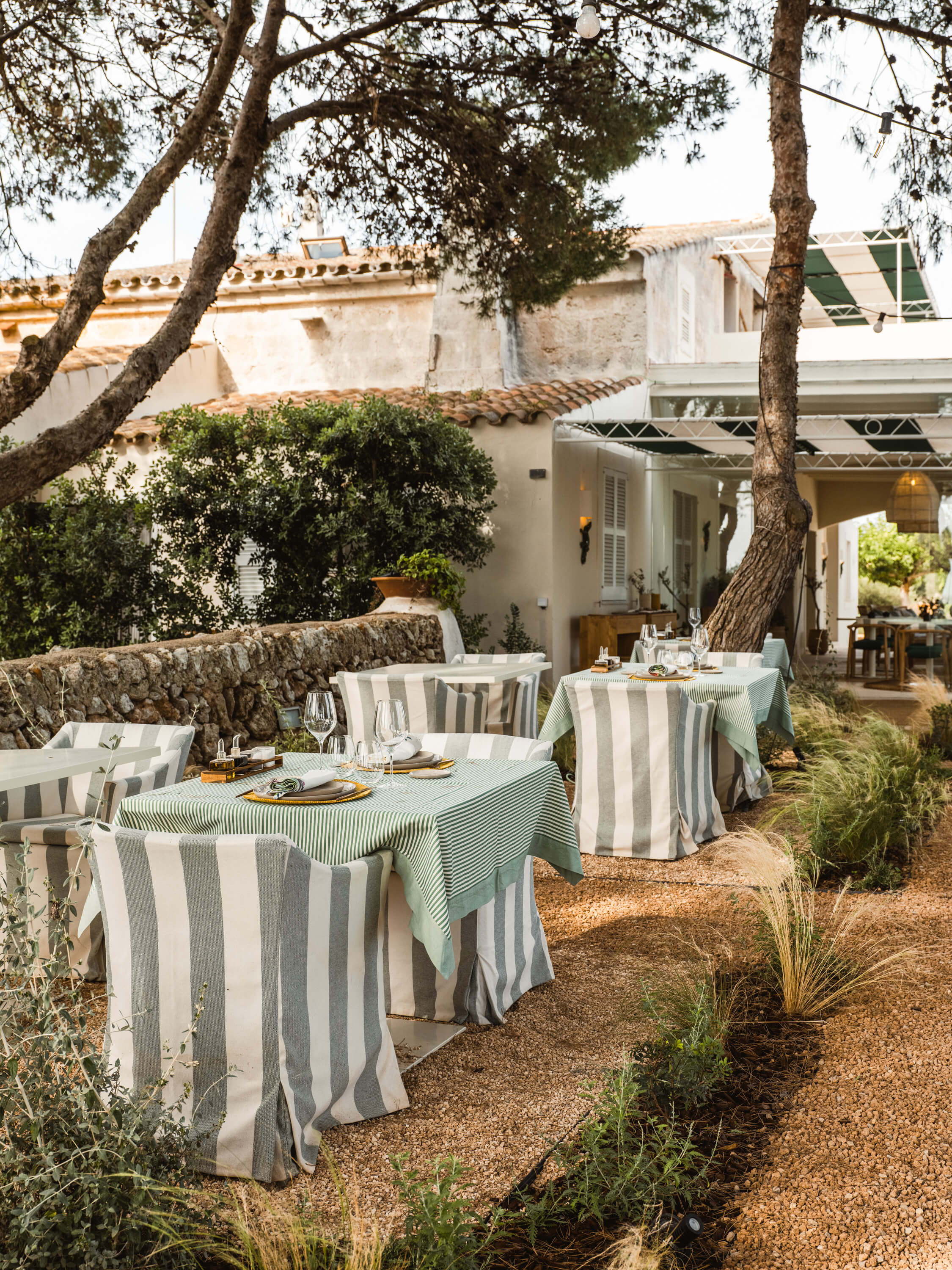 Morvedra Nou, Balearic Islands — Outdoor dining terrace with striped linen tablecloths beneath ancient trees, stone walls, and whitewashed Mediterranean architecture.