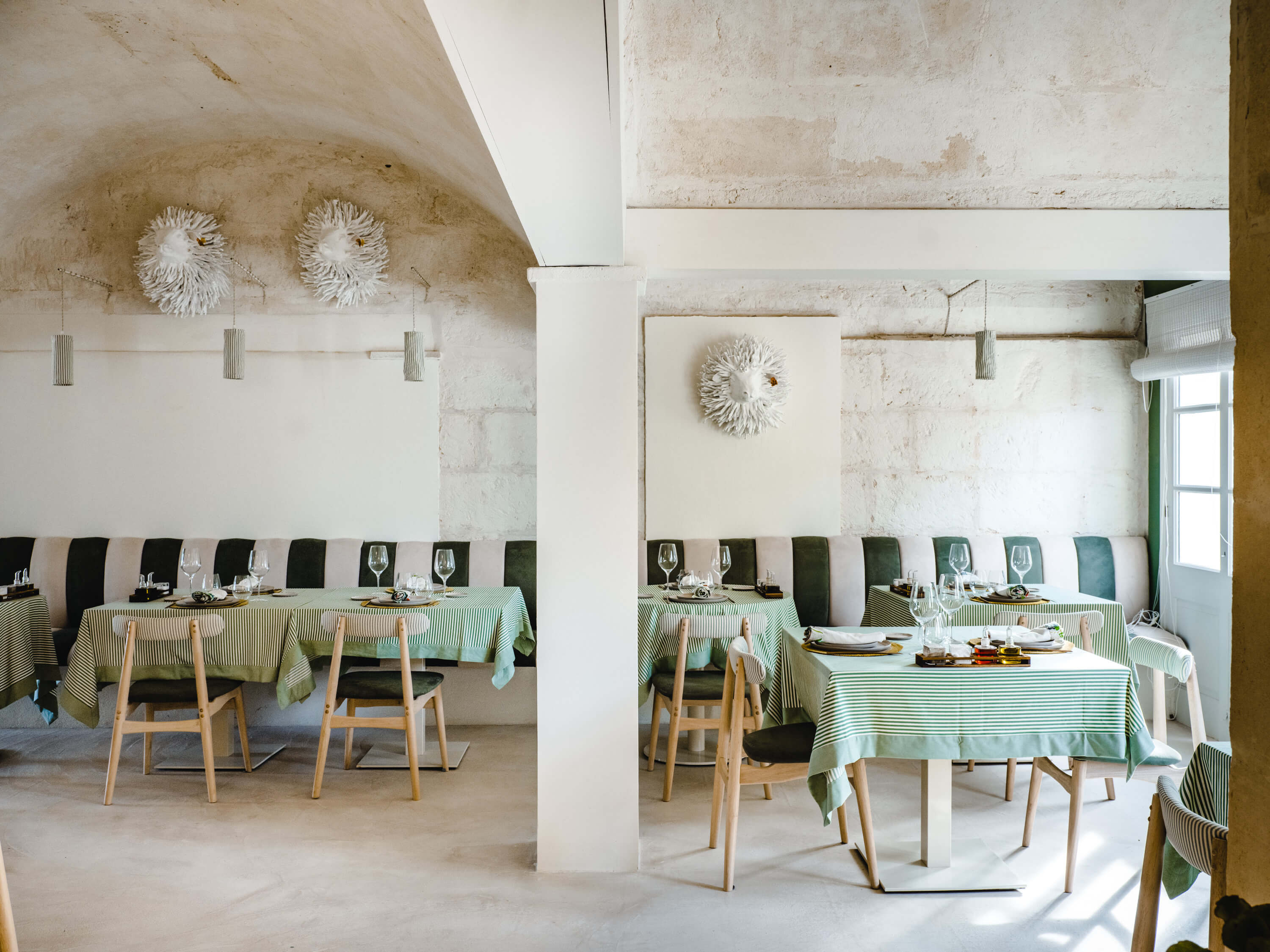 Morvedra Nou, Balearic Islands, Spain — Minimalist dining room with whitewashed stone walls, green-striped linen tablecloths, natural wood chairs, pendant lights, and woven wall sculptures.