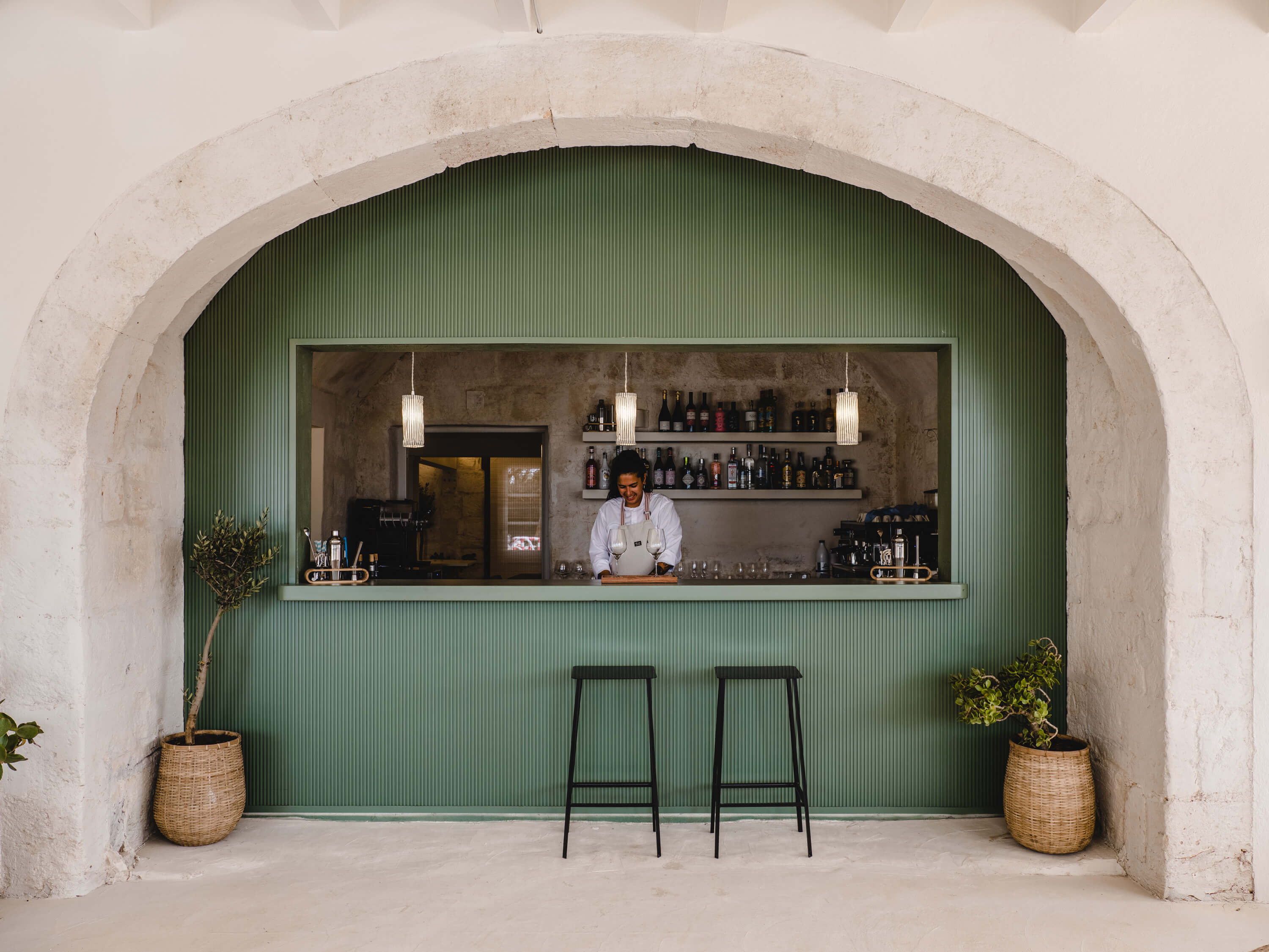 Morvedra Nou, Balearic Islands — Arched stone bar with sage green corrugated panels, pendant lights, dark stools, bottle-lined shelves, and woven planters flanking the counter.