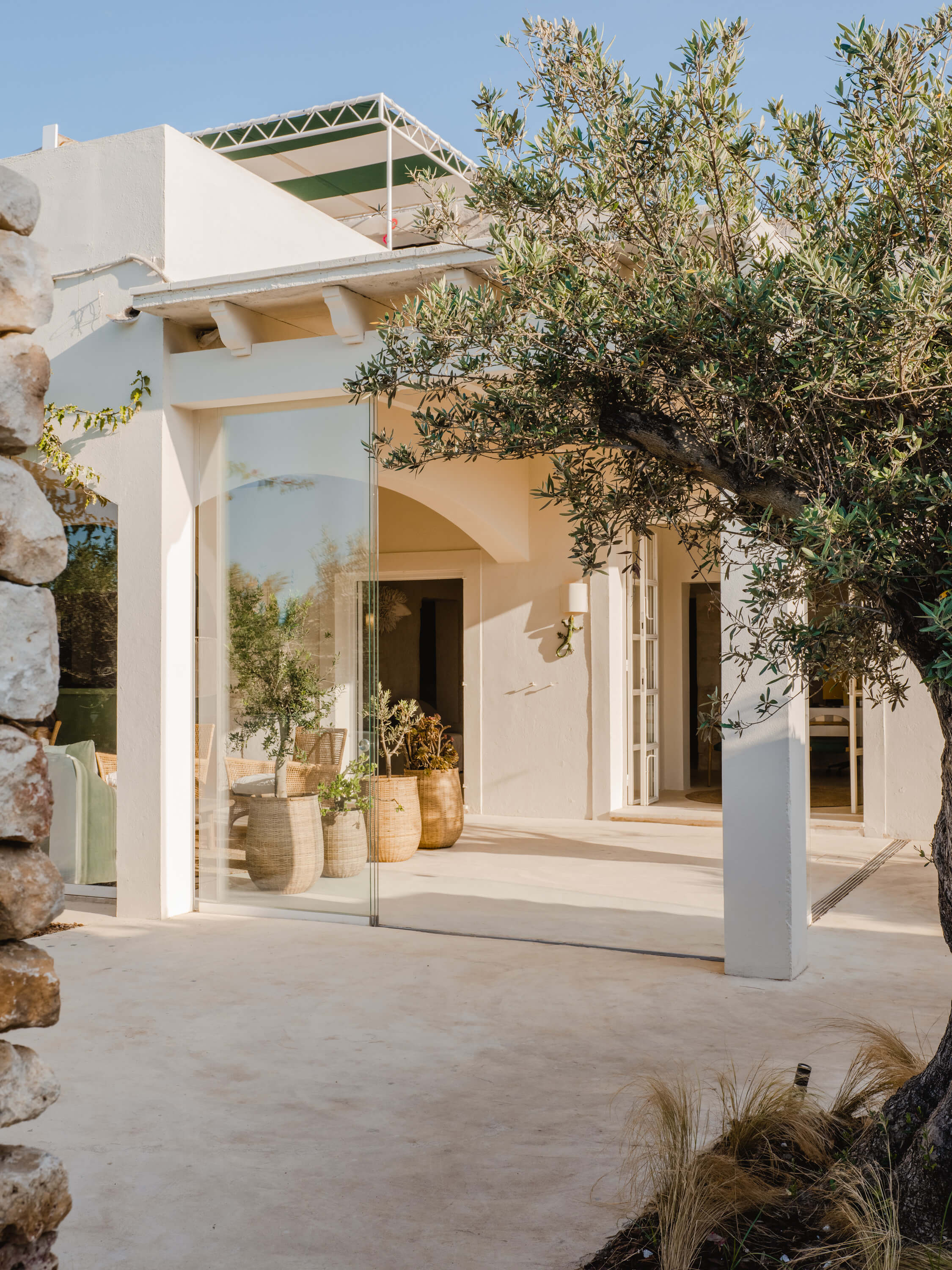 Morvedra Nou, Balearic Islands — Minimalist white pavilion with glass walls, olive trees, woven baskets, and geometric steel pergola casting sharp shadows on limestone terrace.