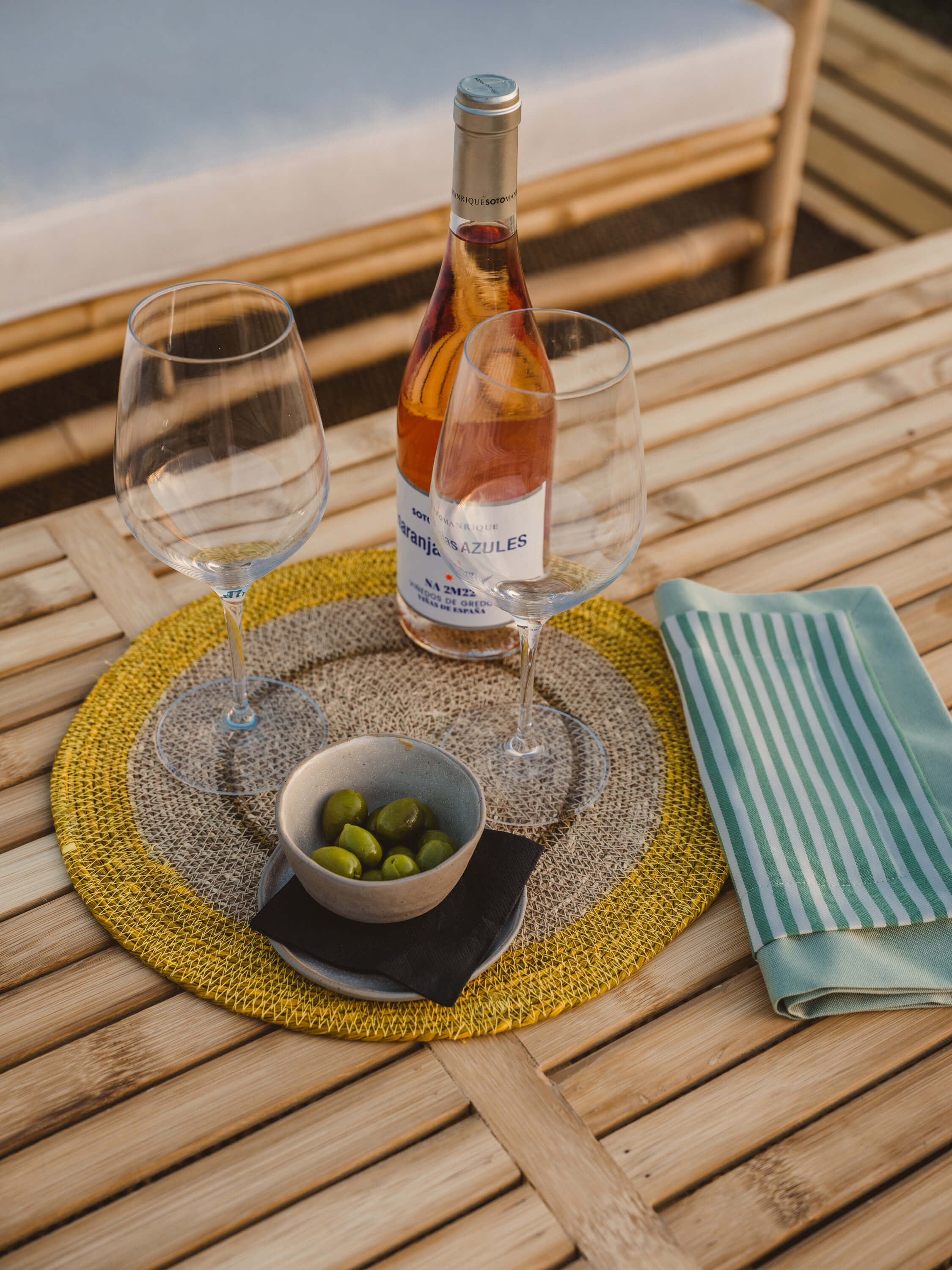 Morvedra Nou, Balearic Islands — Rosé wine and olives on wooden deck with striped napkin and yellow placemats, Mediterranean terrace setting.