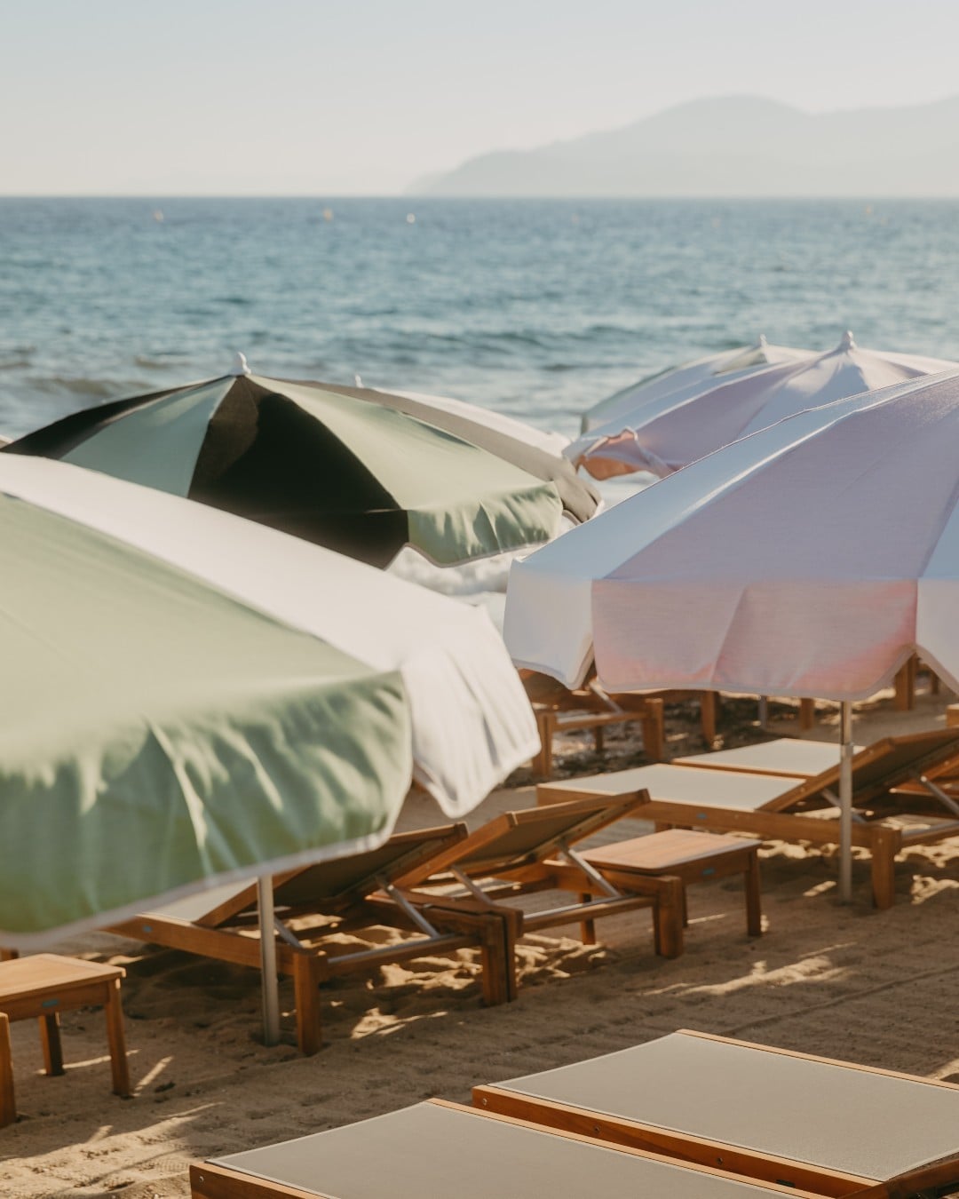 Lily of the Valley, Europe — Wooden loungers with white canvas umbrellas and cushions on sandy beach at sunset, calm sea and distant mountains beyond.