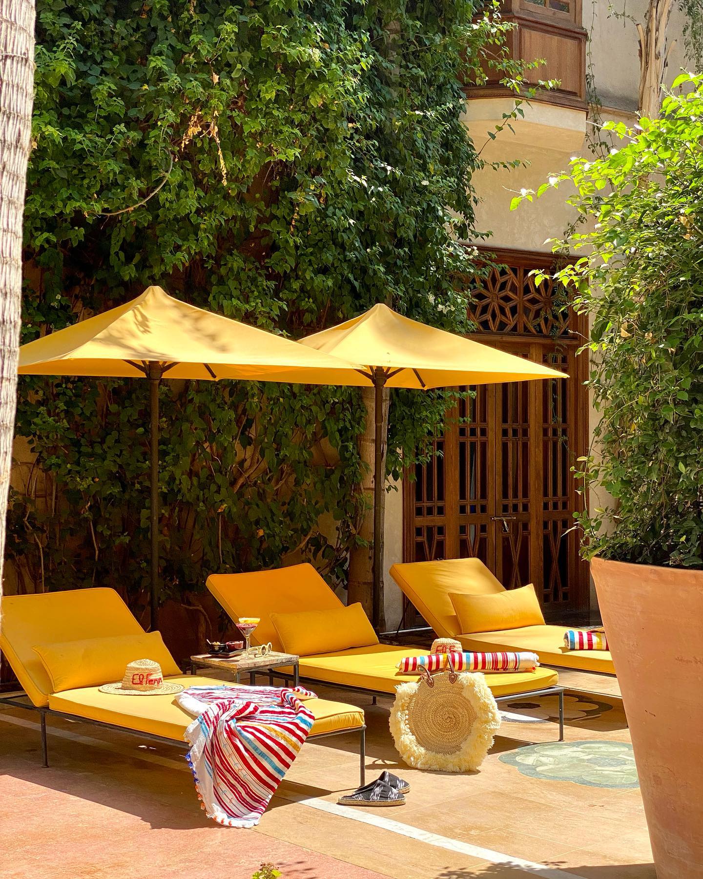 El Fenn, Marrakech, Morocco — Golden loungers beneath striped umbrellas in ivy-draped courtyard with carved wooden doors and terracotta paving.
