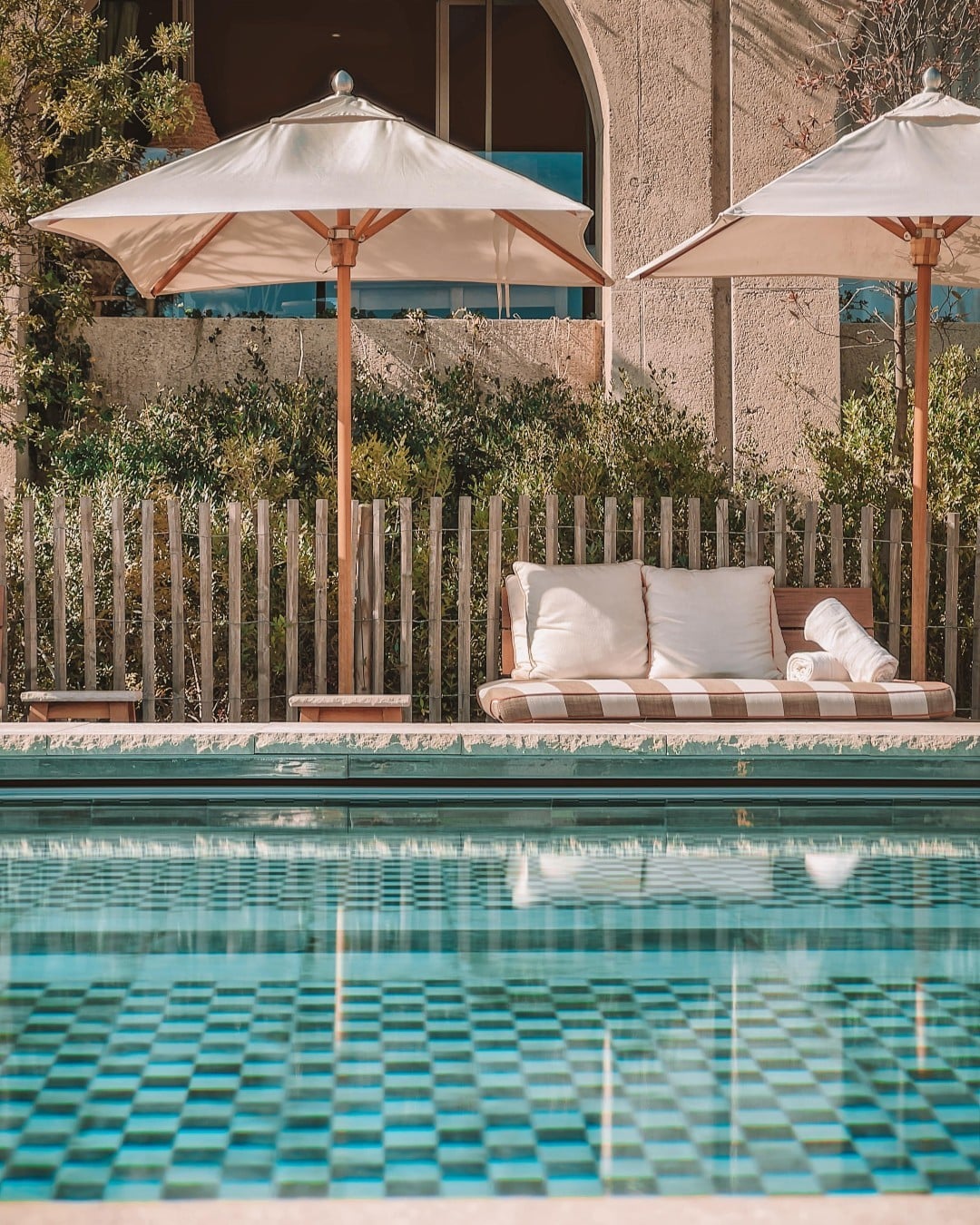 Lily of the Valley, Europe — Poolside lounge with cream umbrellas, striped daybed, wooden fence, and turquoise tiled pool reflecting Mediterranean architecture.