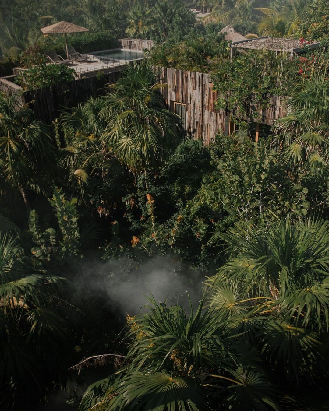 Be Tulum, Mexico — Aerial view of jungle compound with wooden structures, steaming cenote, palm vegetation, and pool deck with umbrella nestled in lush tropical landscape.