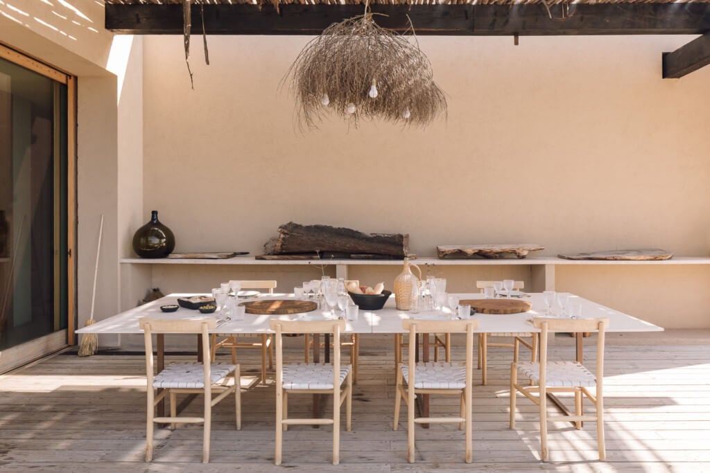Melides Art, Portugal — Minimalist outdoor dining pavilion with whitewashed table, natural wood chairs, thatched pendant light, and ochre stucco walls.