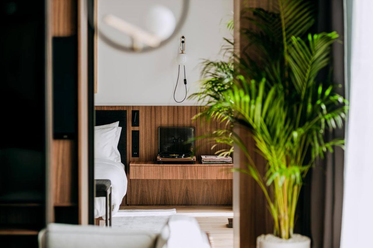 Nobu Warsaw, Poland — Minimalist hotel bedroom with warm wood paneling, white bedding, potted palm plant, and modern fixtures in natural light.