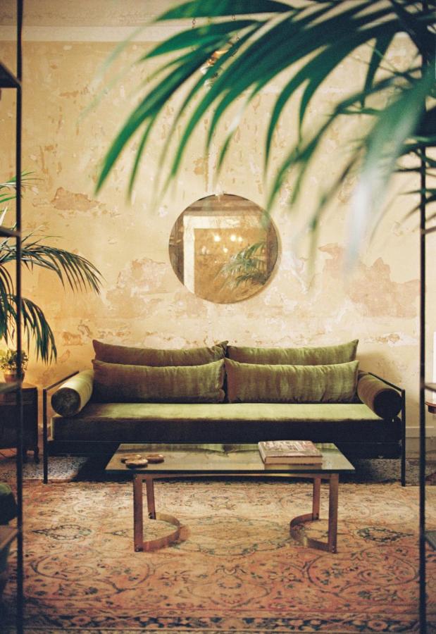 Shila, Europe — Sunlit sitting room with olive linen sofa, round mirror, potted palms, and patterned rug casting botanical shadows on textured walls.