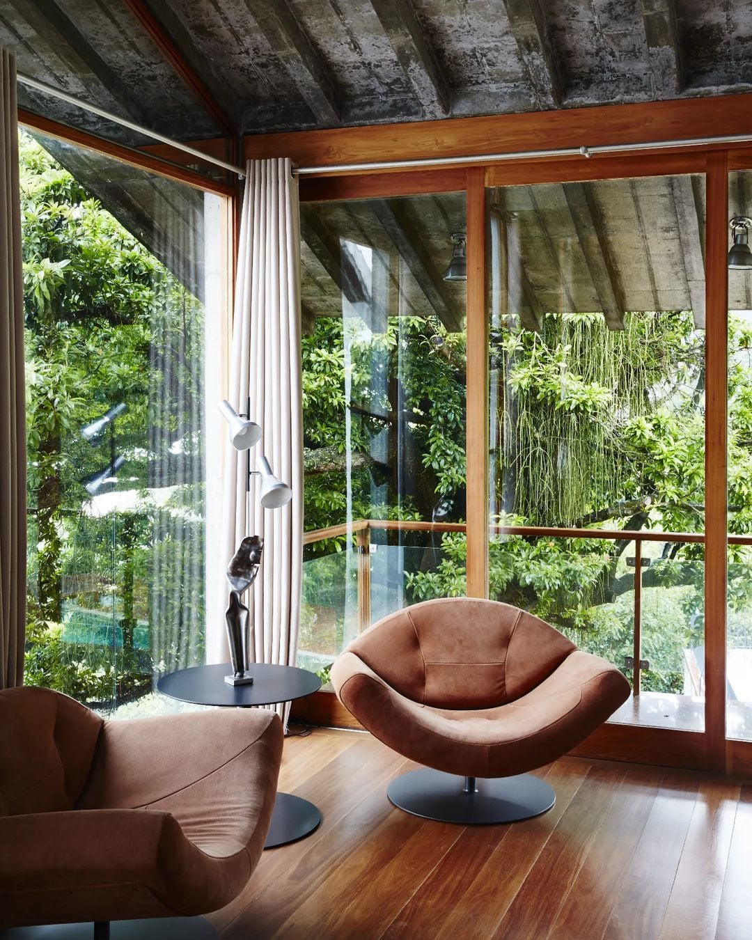 Chez Georges, South America — Mid-century curved leather armchairs with black pedestal bases facing floor-to-ceiling windows overlooking lush green garden, concrete ceiling, warm wood flooring.