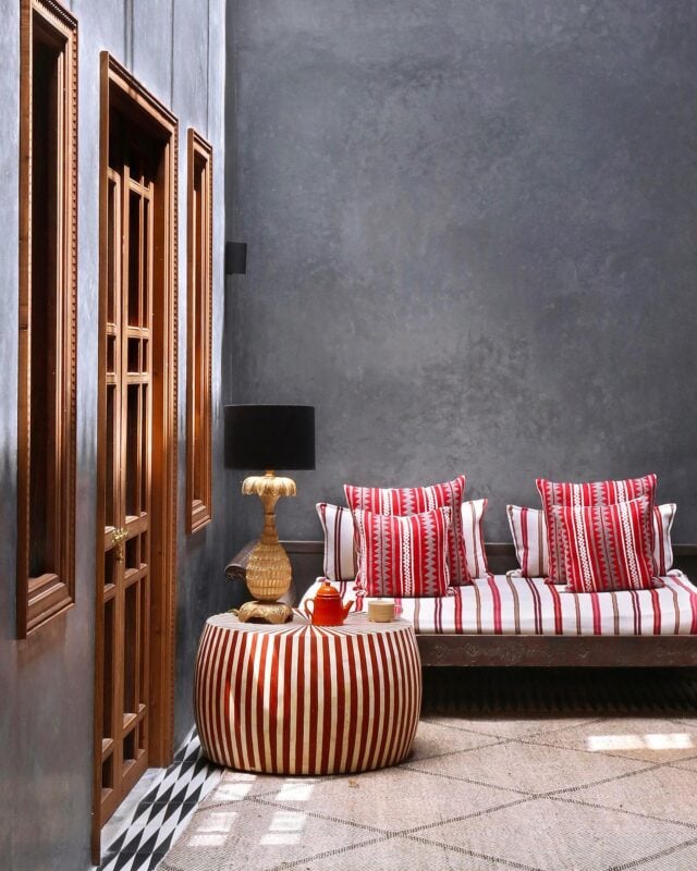 El Fenn, Marrakech, Morocco — Striped red and white daybed with matching pillows beside carved wooden doors and round pouf table with brass lamp, concrete walls and geometric tile floor.