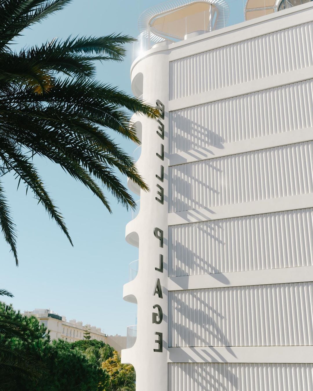 Belle Plage, Europe — Modernist white façade with vertical ribbed panels and curved balconies, palm fronds in foreground, clear sky, coastal luxury architecture.