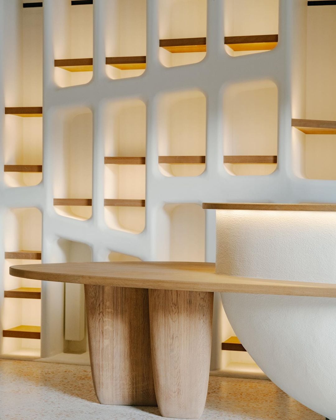 Belle Plage, Europe — Wooden table with curved legs beneath backlit wall niche shelving and warm ambient lighting in minimalist interior.