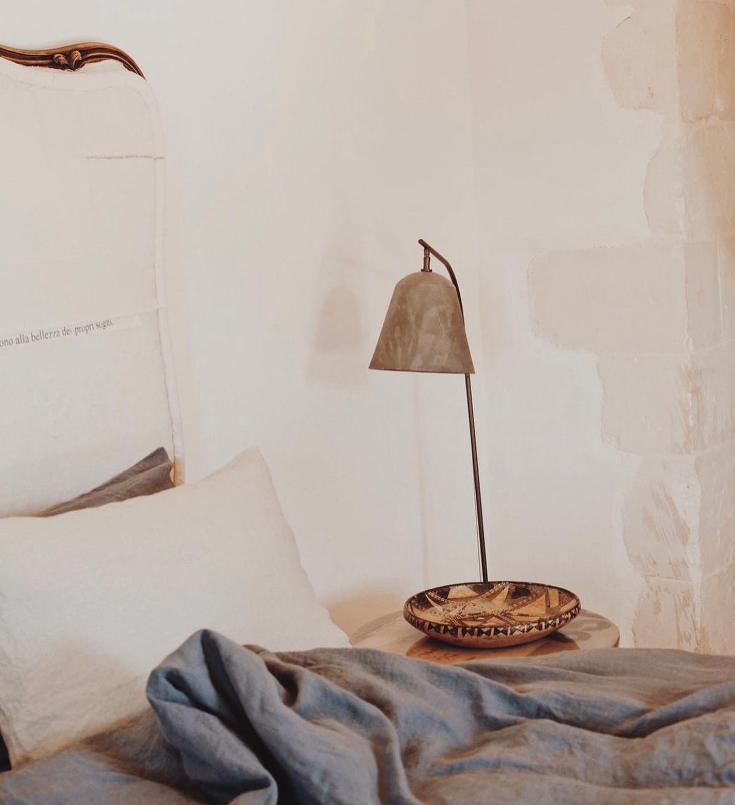 Dimora delle Balze, Europe — Rustic bell suspended above woven basket on linen bed with curved wooden furniture and whitewashed walls.