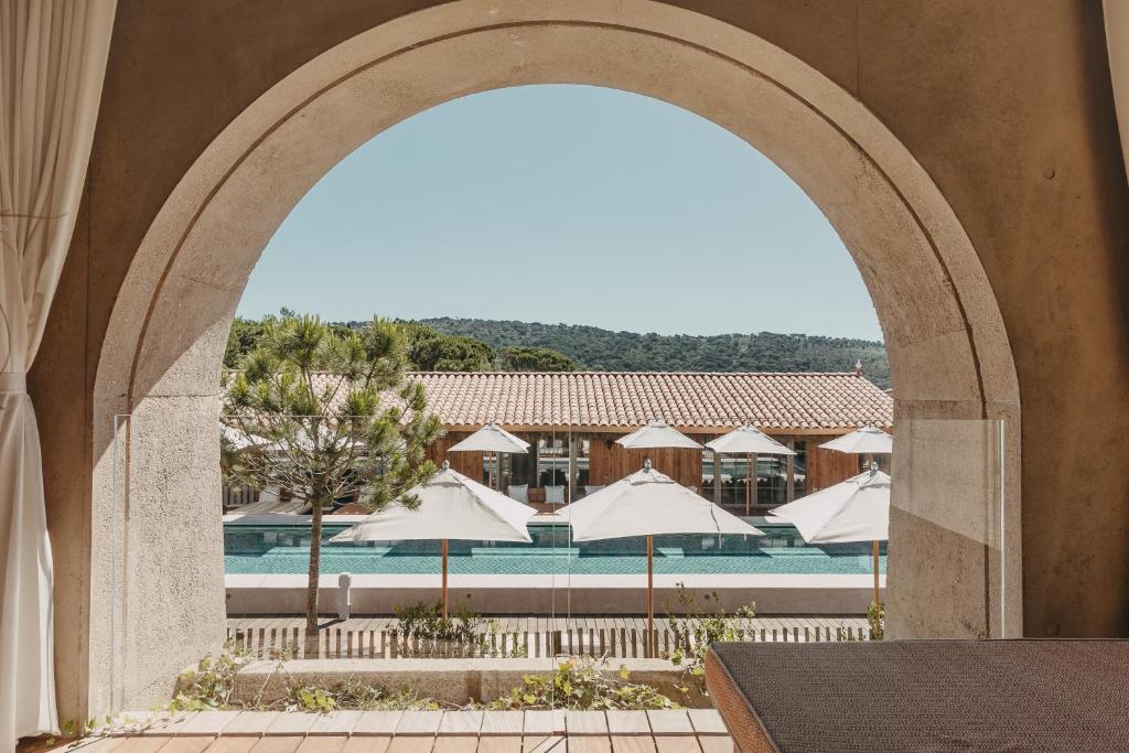 Lily of the Valley, Europe — Arched stone colonnade framing pool terrace with white umbrellas, minimalist wood pavilion, and forested hills under clear sky.