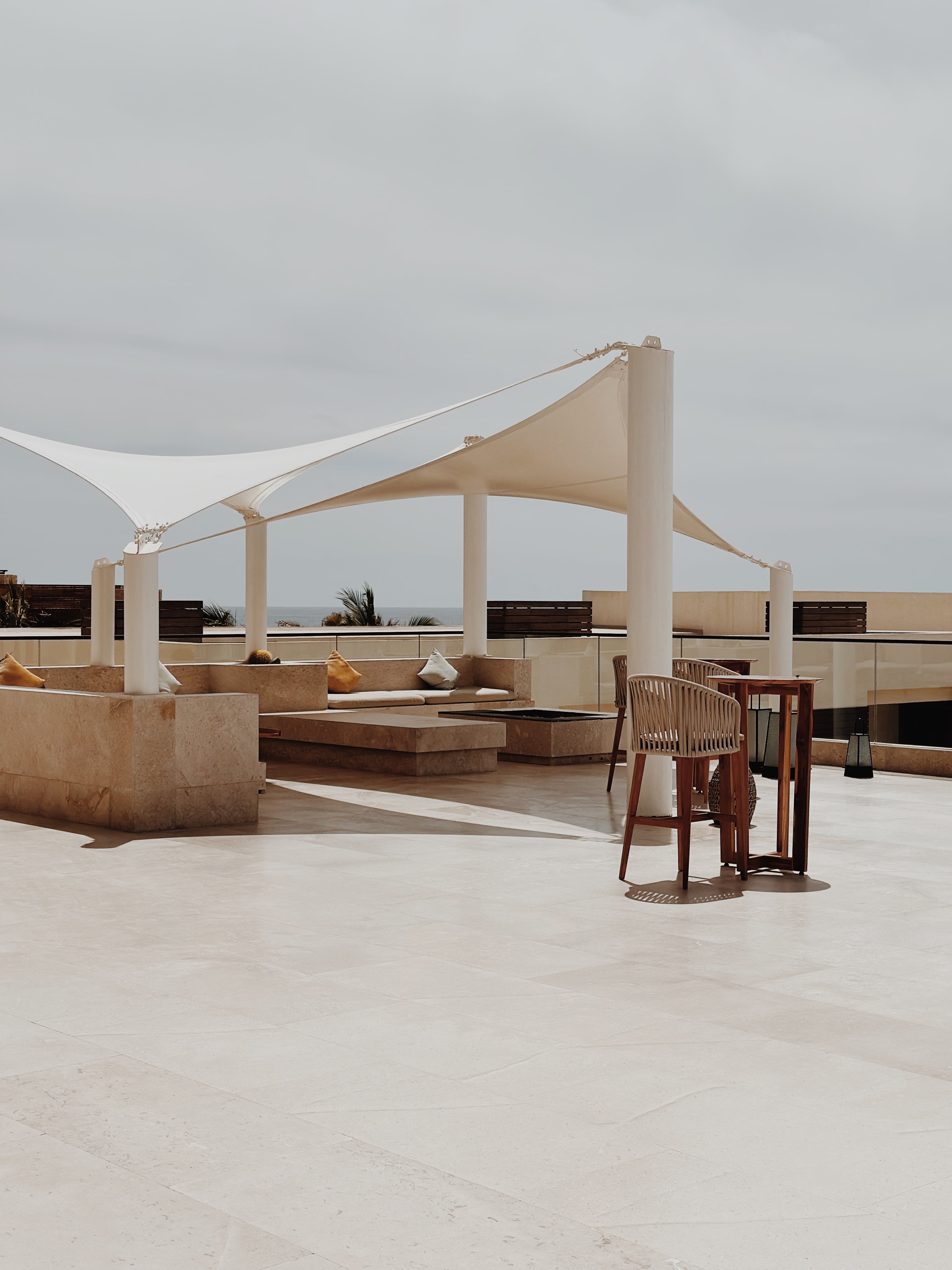 Nobu Los Cabos, Mexico — Minimalist rooftop pavilion with cream sail canopy, concrete seating, wooden barstool, ocean horizon view, overcast sky.