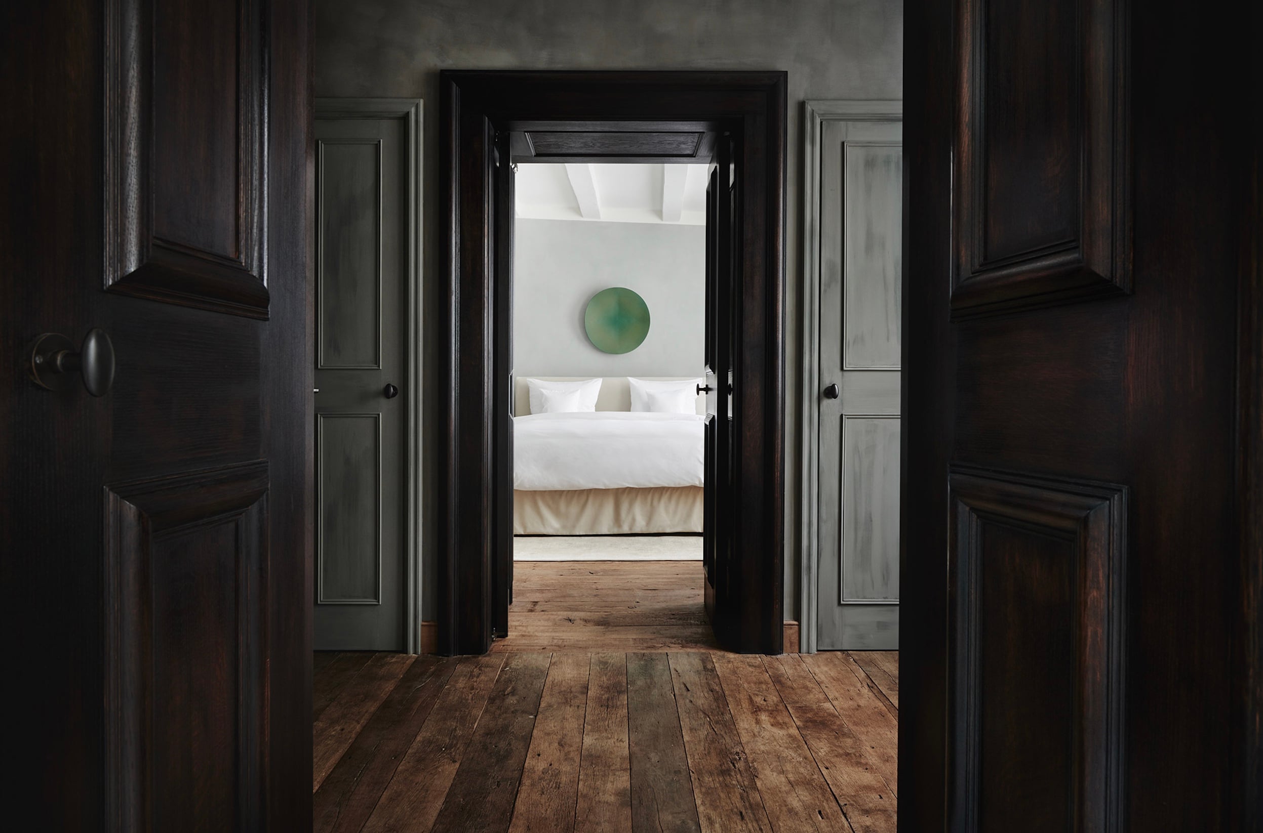 Purs, Europe — Dark timber hallway with black doors opening to minimalist bedroom, white bedding, green circular artwork, natural light from skylight.