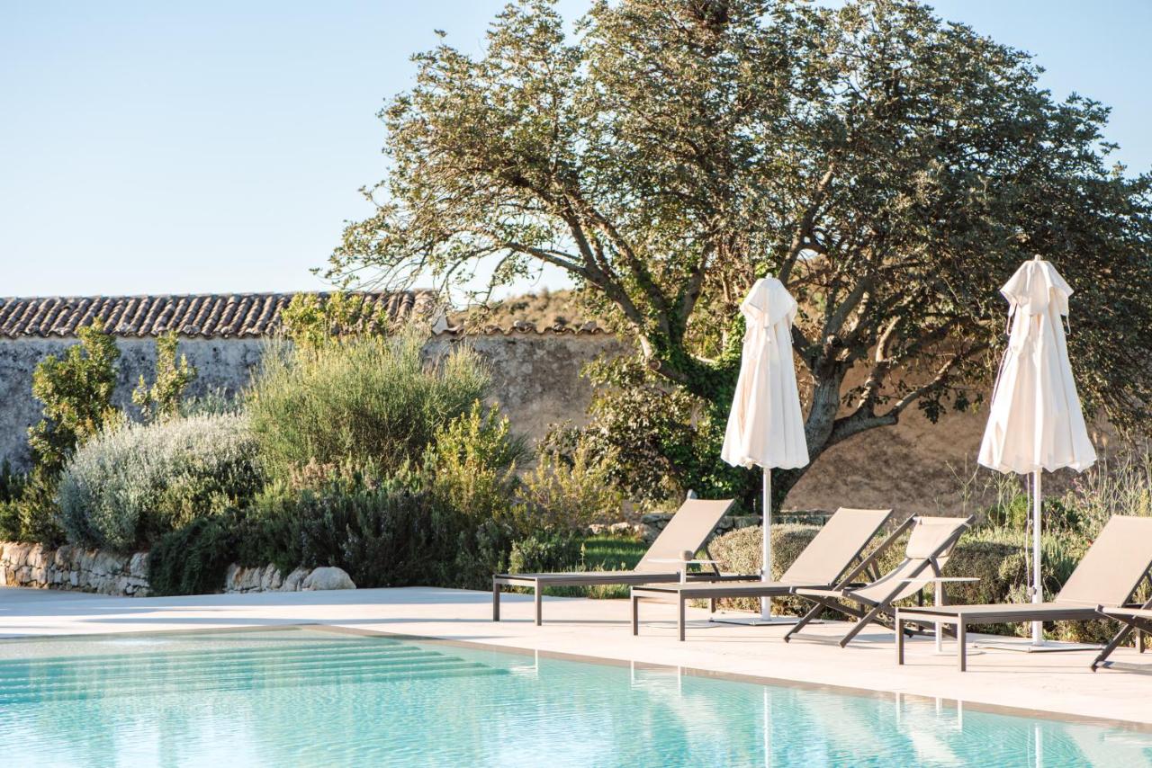 Dimora delle Balze, Europe — Poolside lounge with cream umbrellas, teak loungers, terracotta paving, olive trees, and stone villa architecture in warm Mediterranean light.