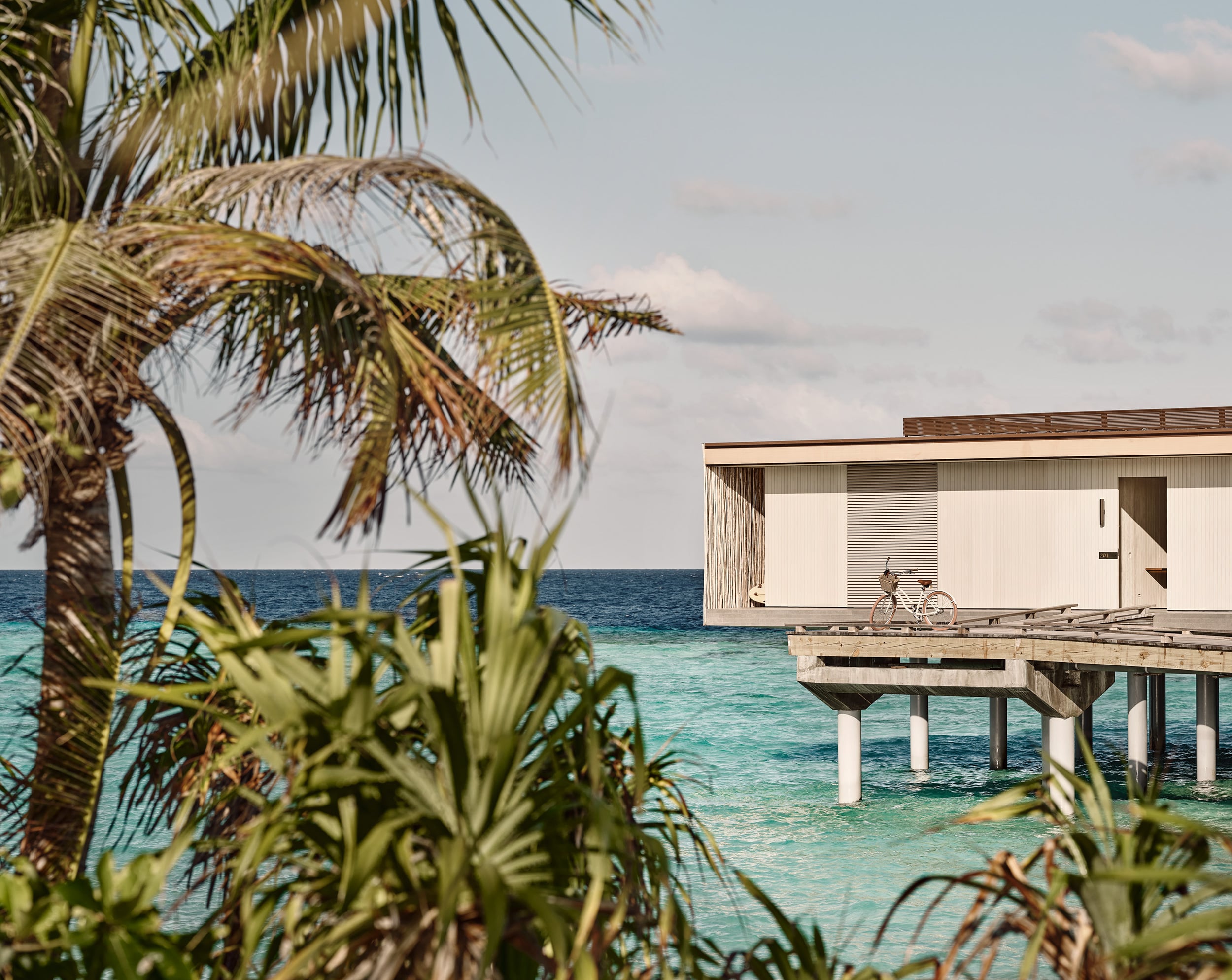 Patina Maldives, Asia — Overwater bungalow with white corrugated exterior and bicycle on wooden deck, turquoise lagoon and palm fronds in foreground.